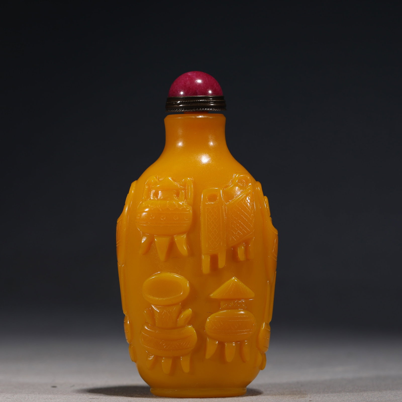 A Chinese Yellow Glass Snuff Bottle Carved with Antique Vessels,Qianlong Mark: A Chinese opaque yellow glass snuff bottle of slender ovoid form,carved in high relief with various antique vessels,including a tripod censer and a vase.It is fitted with a red glass stopper.The base 