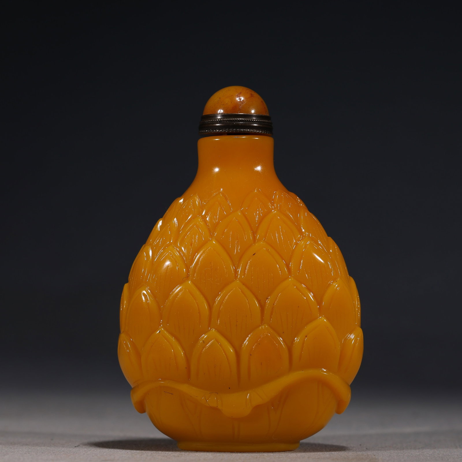 A Chinese Yellow Glass Lotus-Form Snuff Bottle: A Chinese opaque yellow glass snuff bottle carved in the form of a lotus bud,with overlapping petals resting upon a leafy base.It is fitted with an amber-colored stopper.The base is carved with a rais