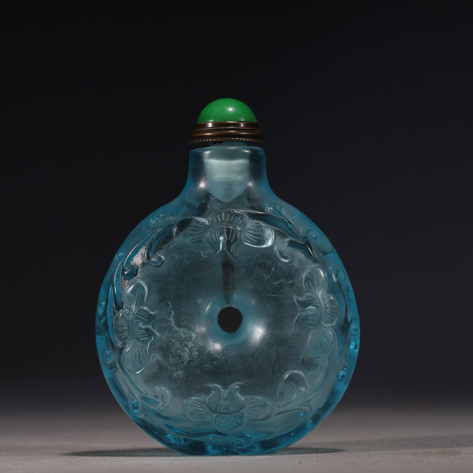 A Chinese Blue Glass Snuff Bottle with Floral Motifs,Qianlong Mark: A translucent blue glass snuff bottle of flattened rounded form,finely carved with continuous floral sprays around a central aperture.It is fitted with a green stopper.The base bears a four-character 