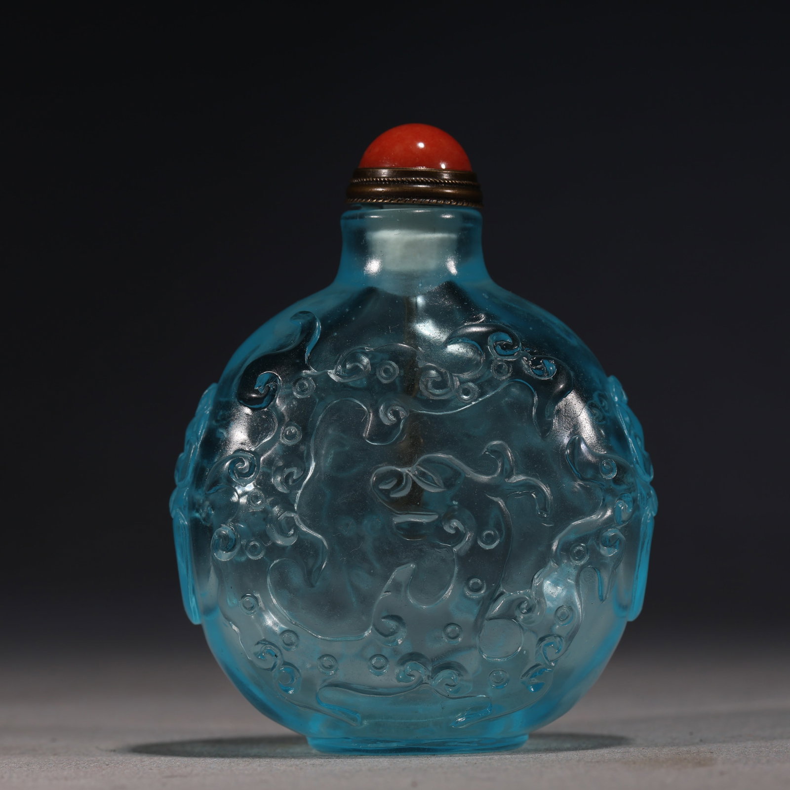 A Chinese Blue Glass Snuff Bottle with Chi Dragons,Qianlong Mark: A translucent blue glass snuff bottle of rounded form,intricately carved in relief with chi dragons and stylized clouds.Fitted with a red glass stopper.The base bears a four-character seal mark readin