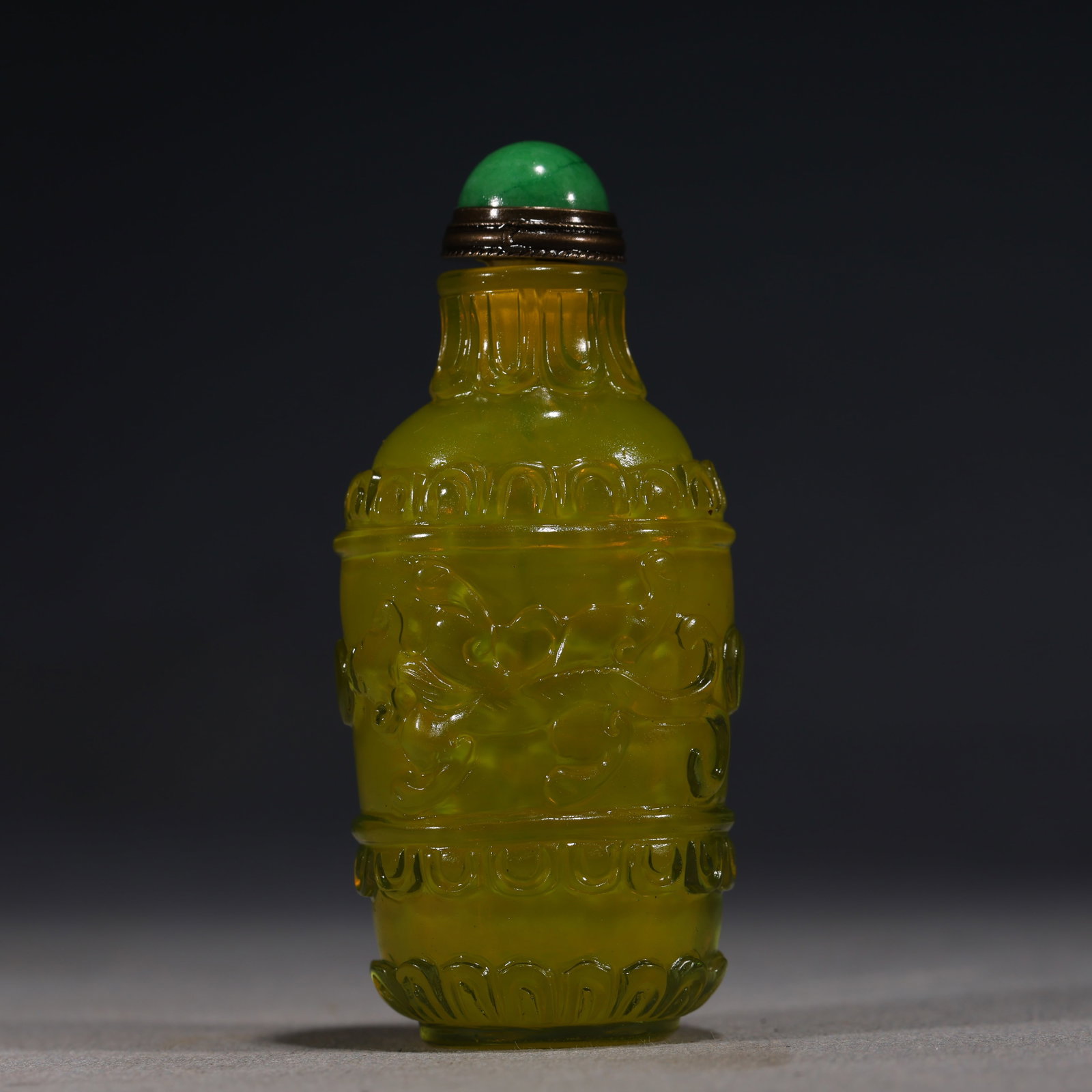 A Chinese Yellow-Green Glass Snuff Bottle with Archaistic Motifs,Qianlong Mark: An opaque yellow-green glass snuff bottle of cylindrical form,carved with stylized archaistic patterns and ruyi heads.Fitted with a green stopper.The base bears a four-character seal mark reading(Qian