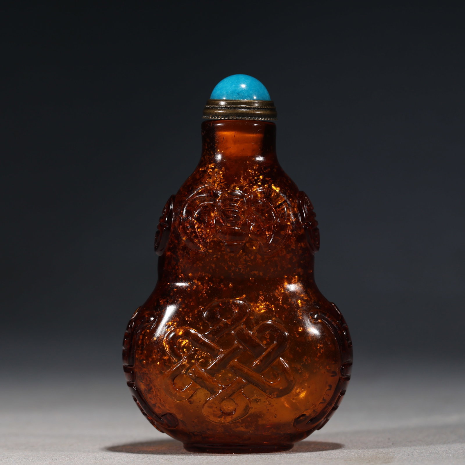 A Chinese Amber-Tone Gold-Speckled Glass Double-Gourd Snuff Bottle,Qianlong Mark: A translucent amber-toned glass snuff bottle suffused with gold speckles,carved in double-gourd form with shou characters and endless knots.Fitted with a turquoise stopper.The base bears a