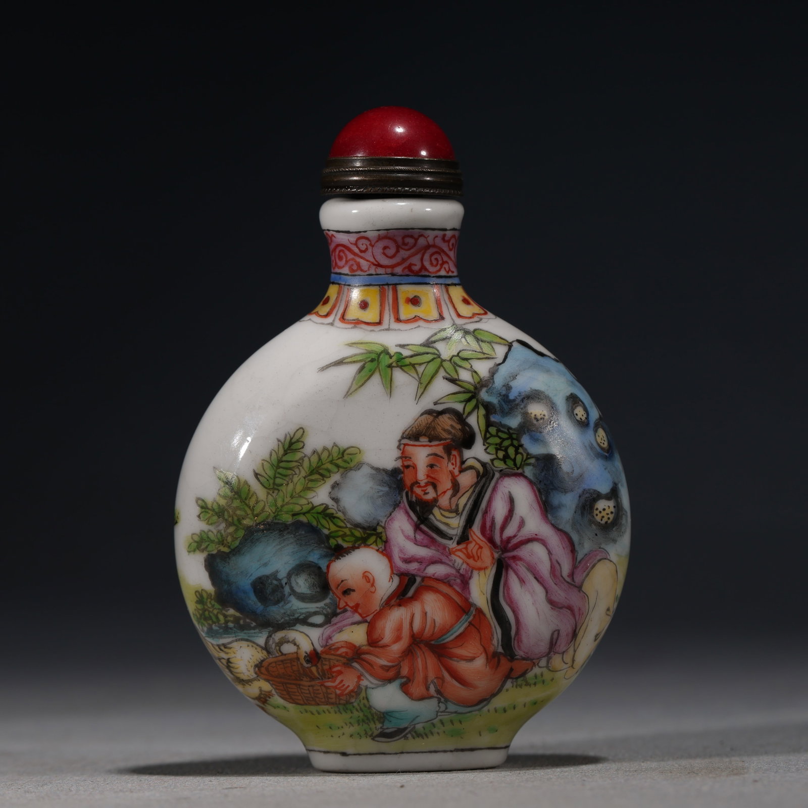 A Chinese Famille Rose Porcelain Snuff Bottle with Figures,Kangxi Mark: A white porcelain snuff bottle decorated in famille rose enamels with a scholar and a boy seated amidst bamboo and rocks.Fitted with a red stopper.The base bears a four-character mark in underglaze bl