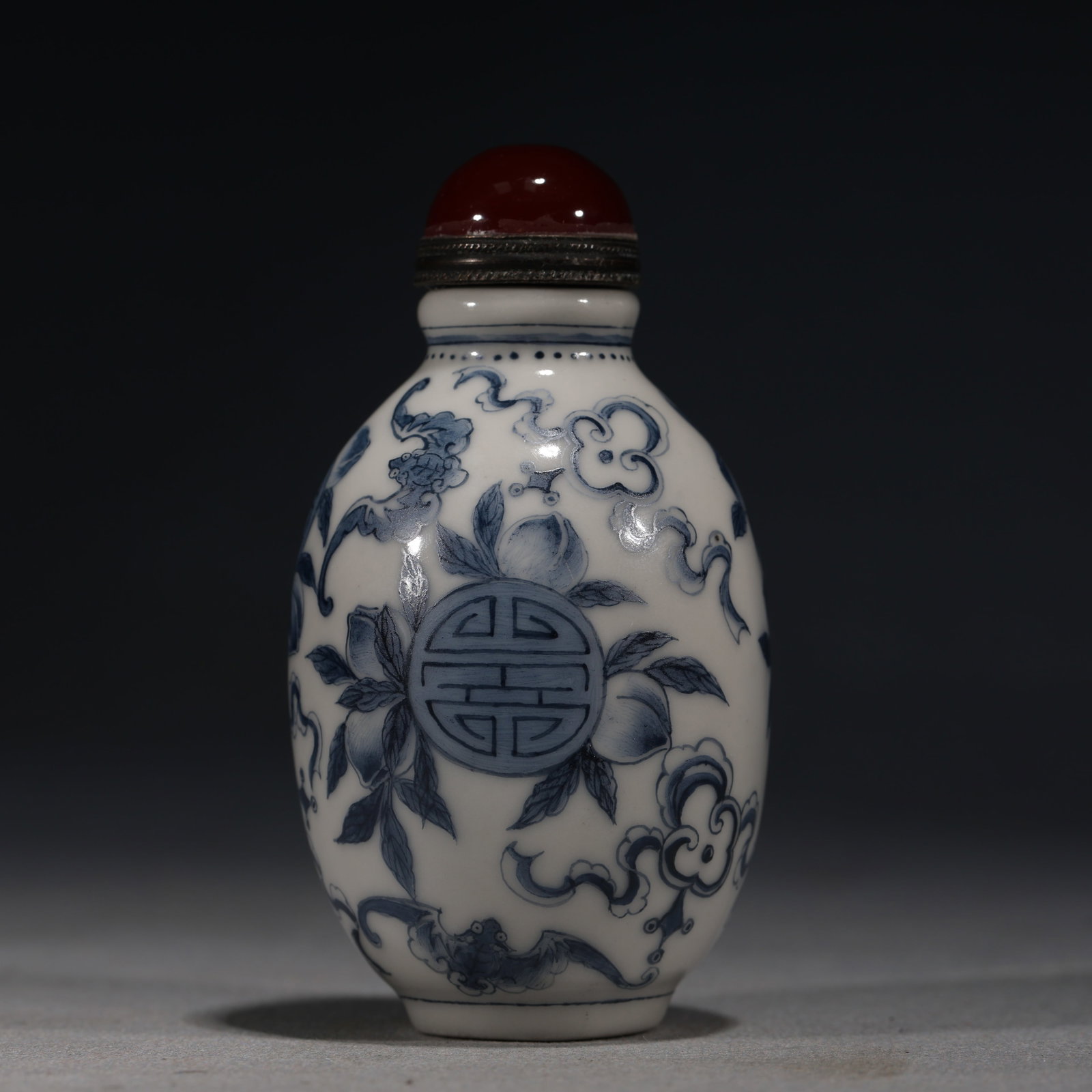 A Chinese Blue and White Porcelain Snuff Bottle with Peaches,Kangxi Mark: A blue and white porcelain snuff bottle of ovoid form,painted with fruiting peach branches,ruyi clouds,and bats surrounding a central shou medallion.Fitted with a red stopper.The base bears a four-cha