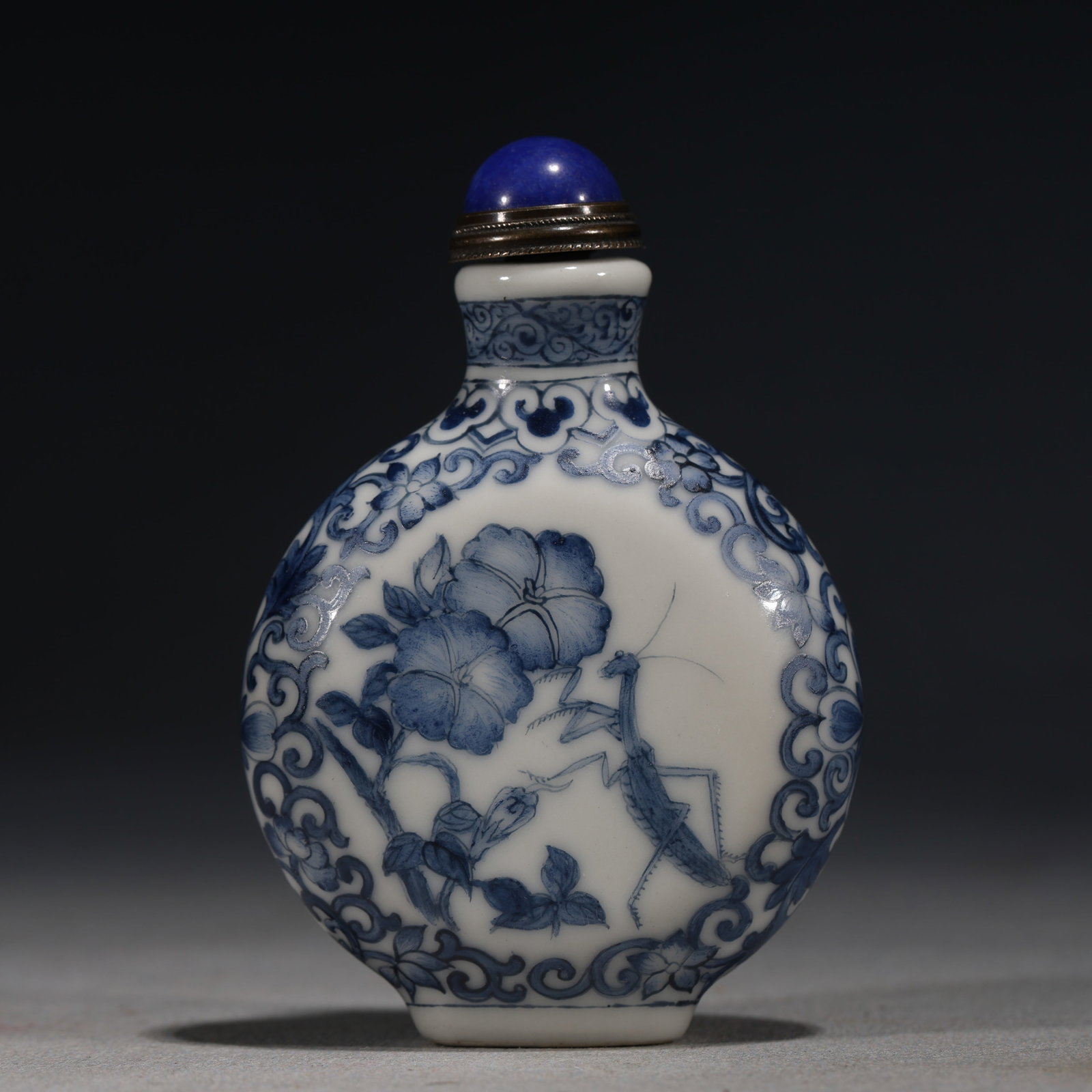 A Chinese Blue and White Porcelain Snuff Bottle with Insects,Qianlong Mark: A blue and white porcelain snuff bottle of rounded form,painted with a continuous floral scroll border enclosing a garden scene of a flower and a praying mantis-insect amidst rocks.Fitted with a lapis