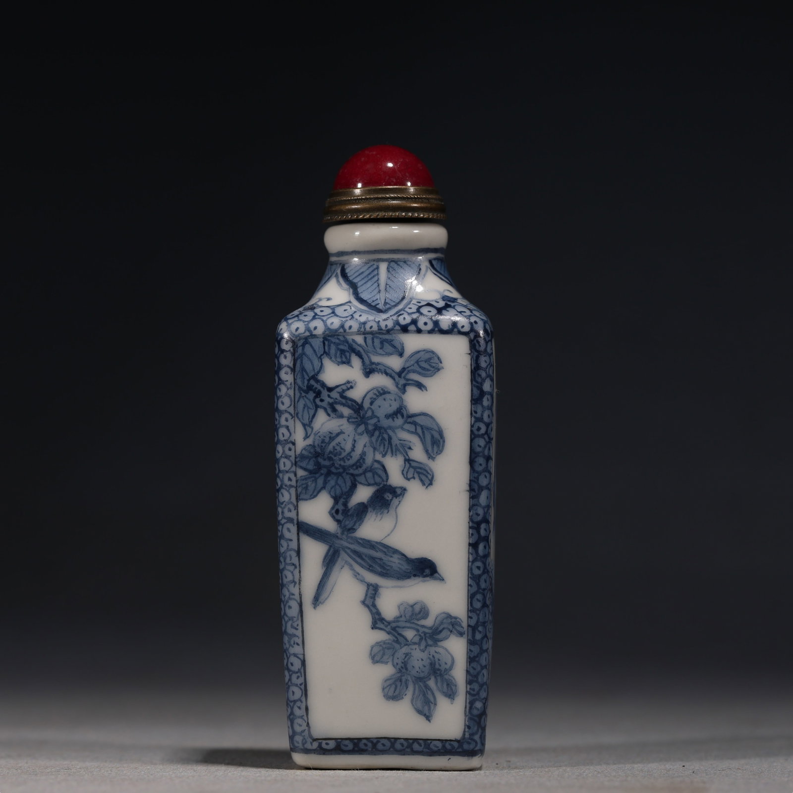 A Chinese Blue and White Porcelain Square Snuff Bottle with Birds and Landscape,Yongzheng Mark: A blue and white porcelain snuff bottle of square form,painted with a bird perched on a flowering branch on one face and a mountain landscape on the other.Fitted with a red glass stopper.The base bear