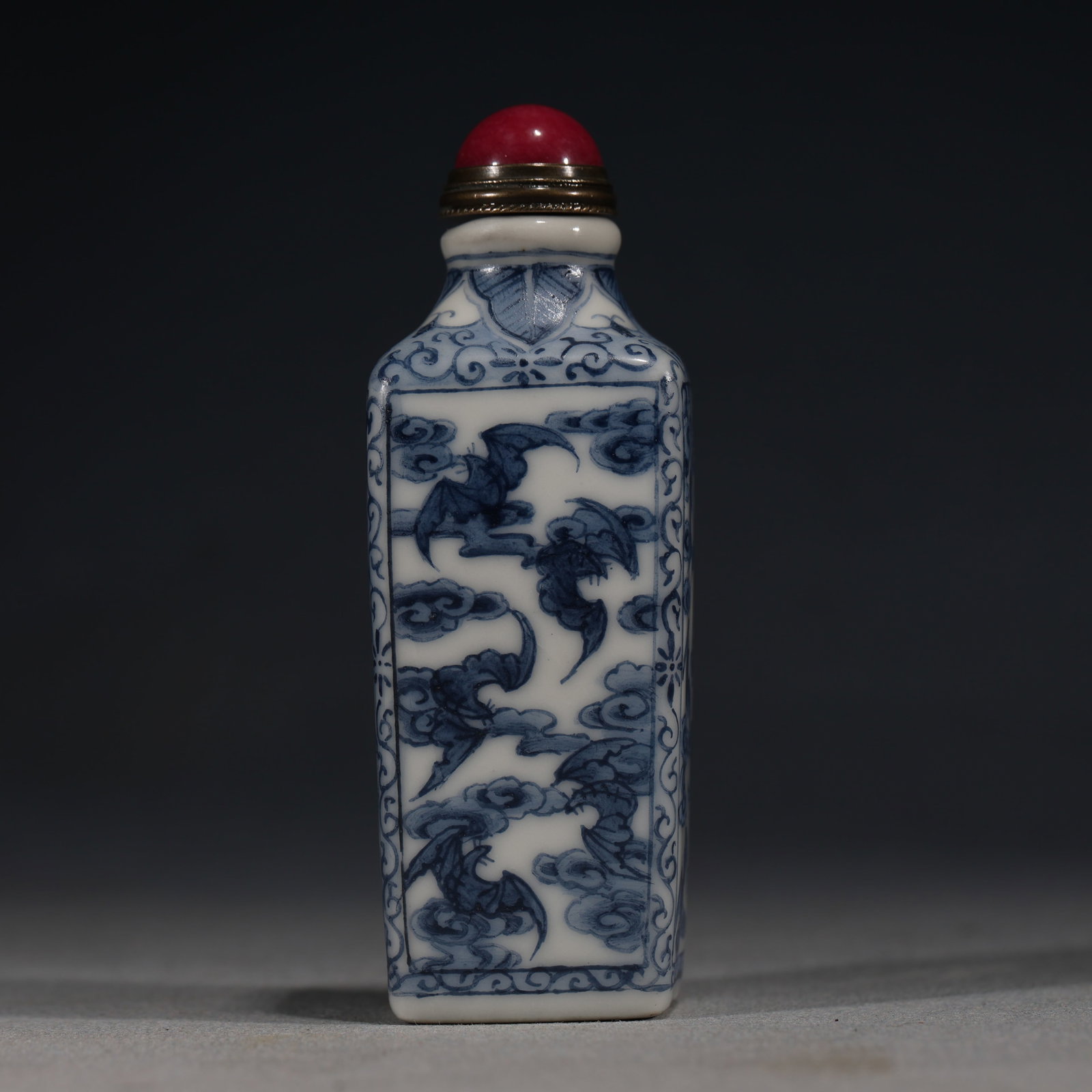 A Chinese Blue and White Porcelain Square Snuff Bottle with Bats and Clouds,Qianlong Mark: A blue and white porcelain snuff bottle of square form,painted with stylized bats flying amidst scrolling clouds.Fitted with a red glass stopper.The base bears a four-character seal mark reading(Qianl