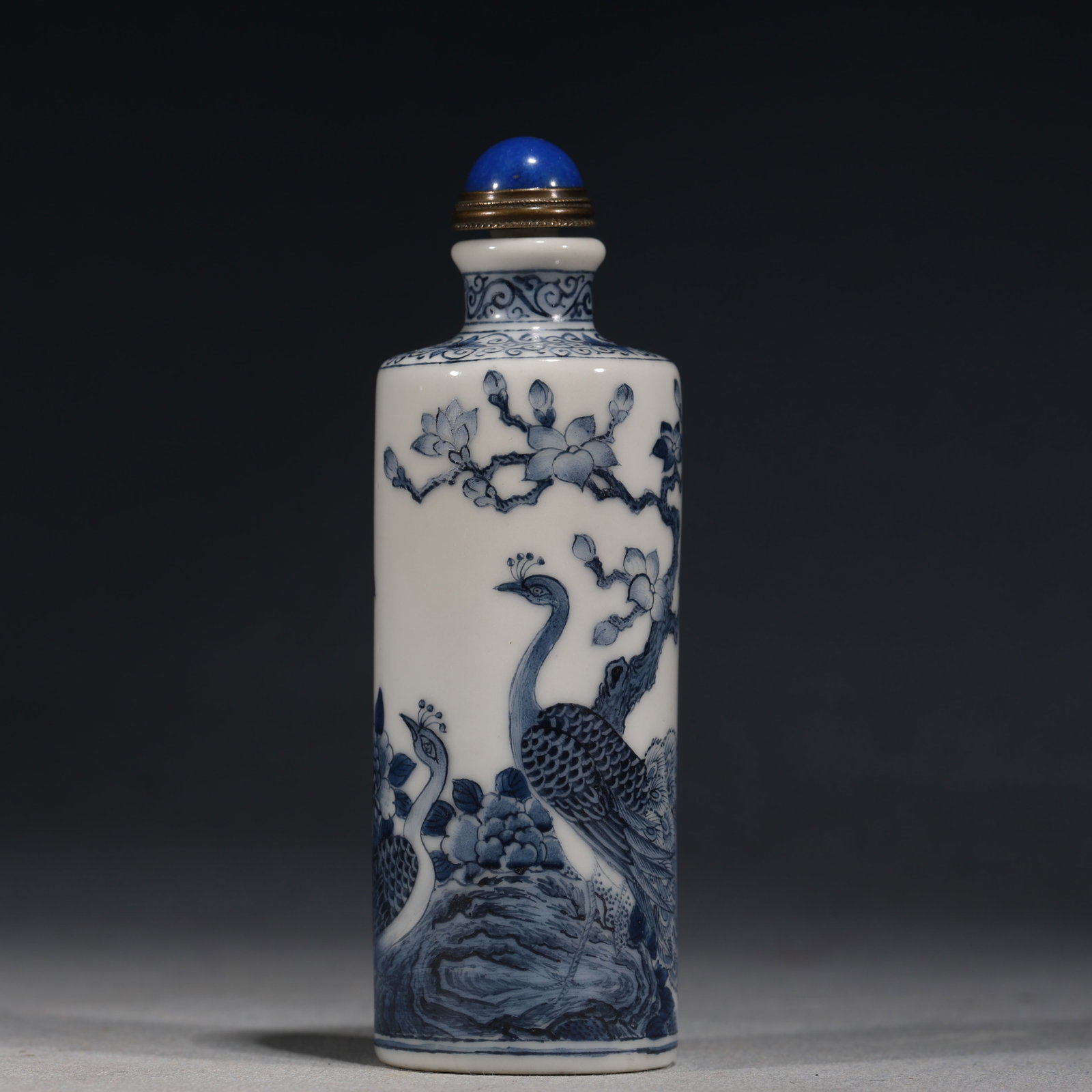 A Chinese Blue and White Porcelain Cylindrical Snuff Bottle with Peacocks and Peonies,Qianlong Mark: A blue and white porcelain snuff bottle of cylindrical form,painted with two peacocks perched on rockwork amidst peony blossoms.Fitted with a blue glass stopper.The base bears a four-character seal ma