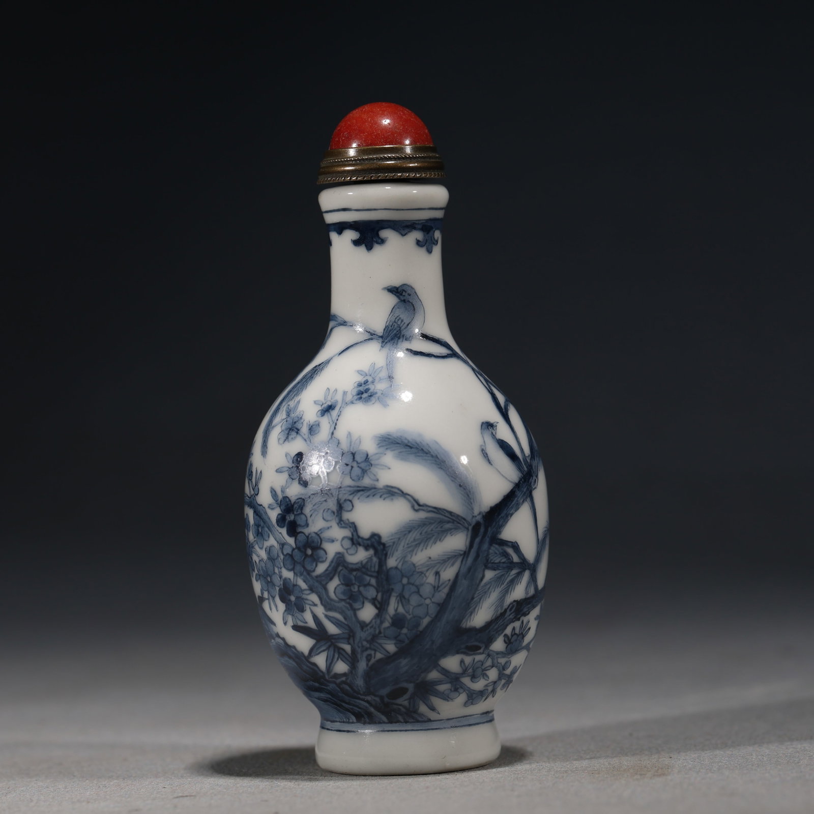 A Chinese Blue and White Porcelain Ovoid Snuff Bottle with Birds,Bamboo and Prunus,Kangxi Mark: A blue and white porcelain snuff bottle of ovoid form,painted with birds perched on branches of pine,bamboo and prunus.Fitted with a red glass stopper.The base bears a four-character regular script ma