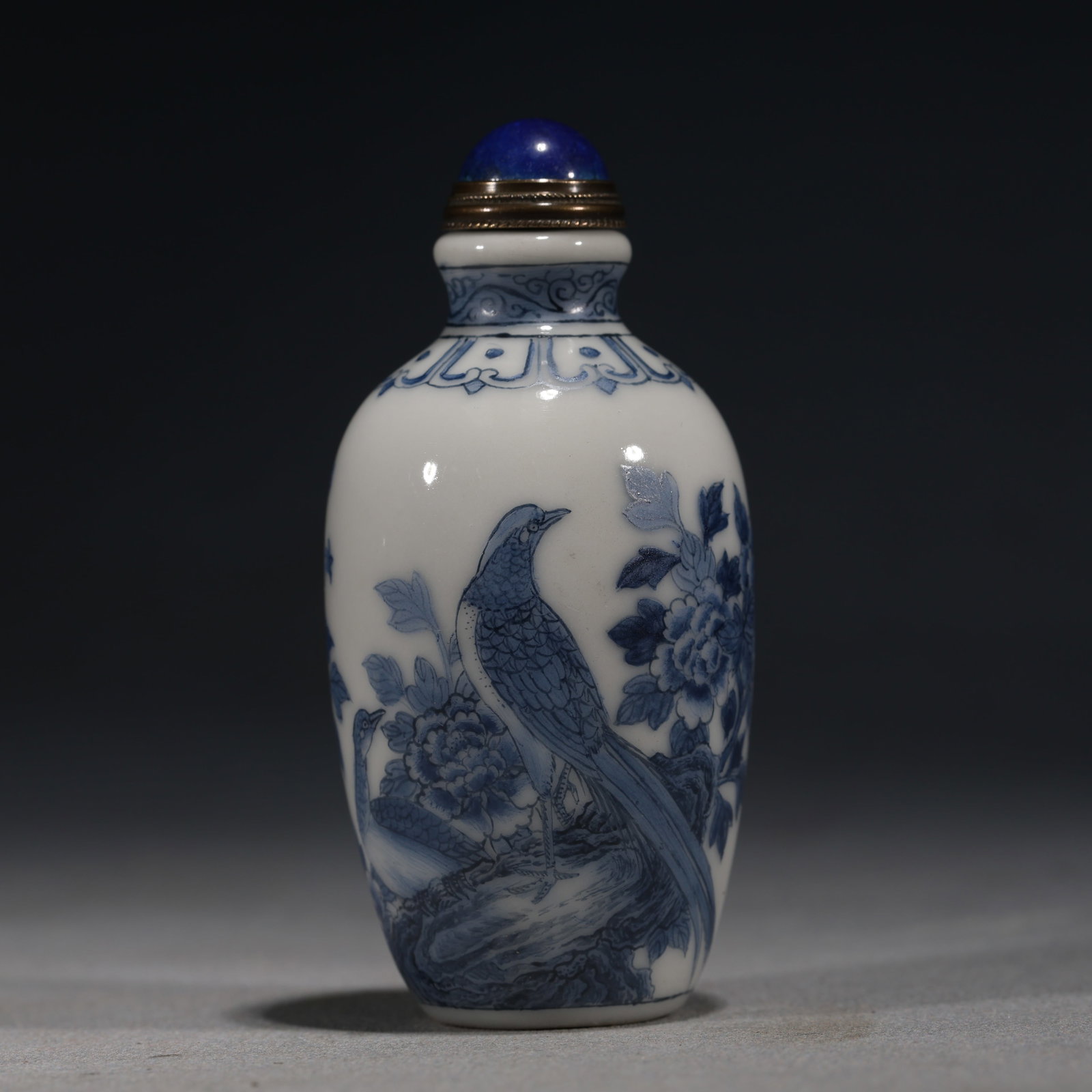 A Chinese Blue and White Porcelain Ovoid Snuff Bottle with Peacock and Peonies,Qianlong Mark: A blue and white porcelain snuff bottle of ovoid form,painted with a long-tailed peacock standing on rockwork beside blooming peonies.Fitted with a blue glass stopper.The base bears a four-character