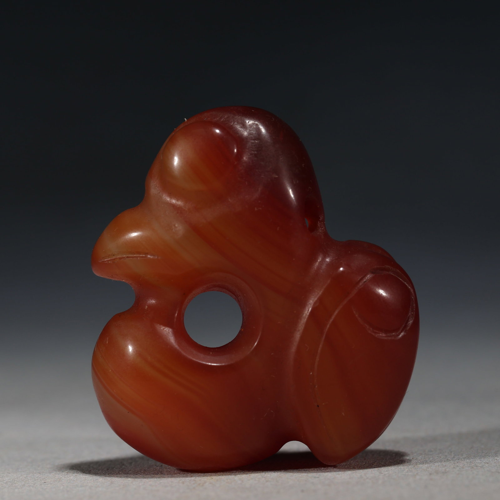 RED AGATE CARVED MYTHICAL BIRD PENDANT: A small,rounded red agate pendant carved in the form of a stylized mythical bird with an arched head,a central circular piercing,and smooth body contours.The stone is polished to a high lustrous finis