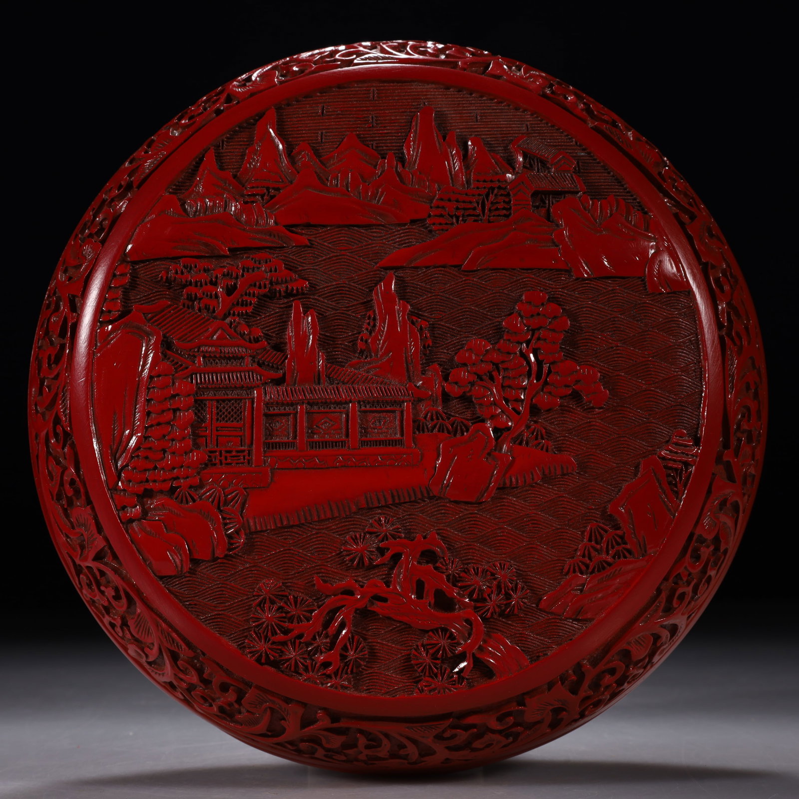 A Carved Cinnabar Lacquer Circular Box with Landscape and Pavilion: Of circular form, the cover presents an expansive landscape with a pavilion built by the water in the center, towering peaks in the distance, and a scholar boating on the rippling water. Period: Qing 