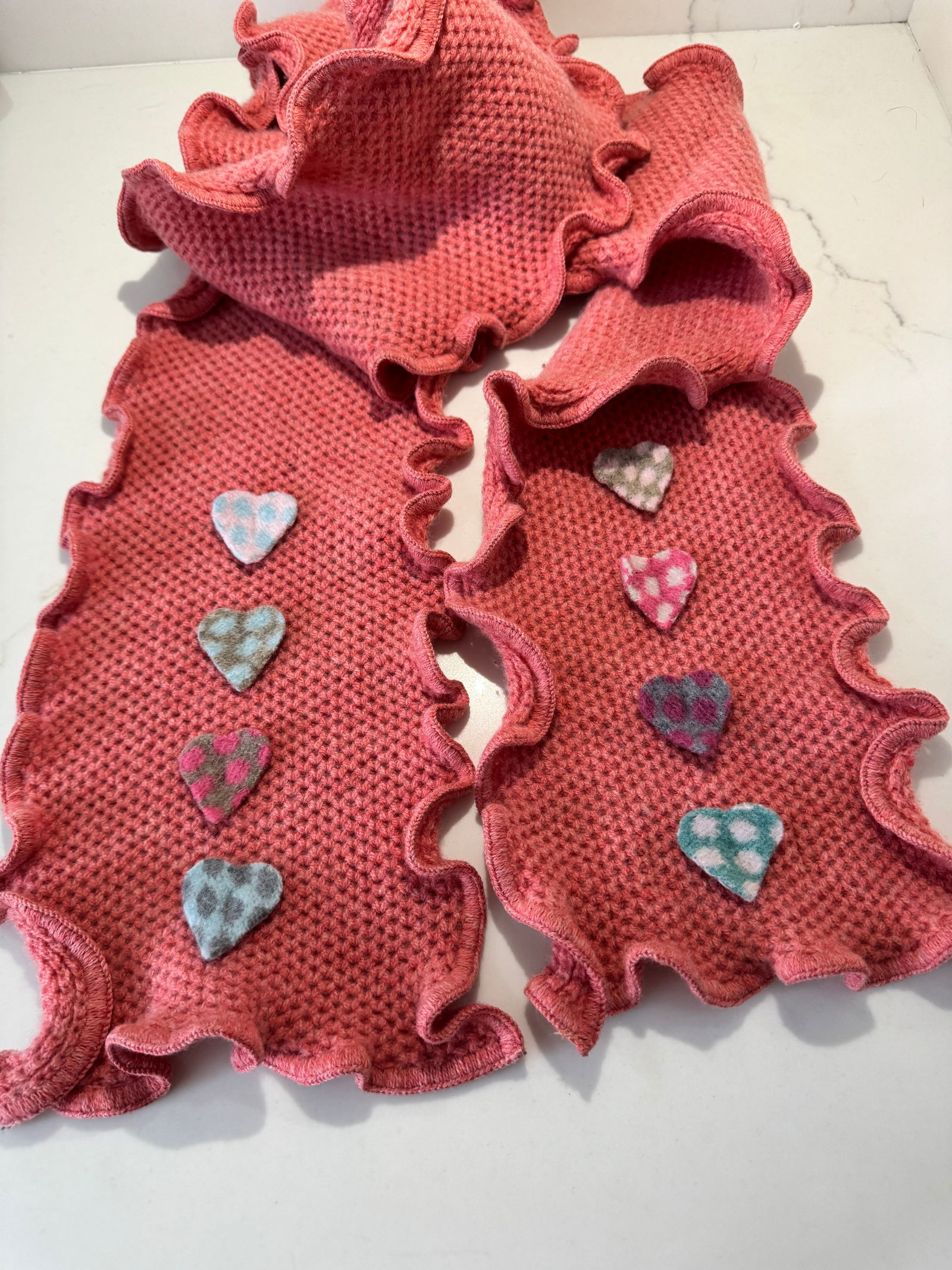 Nicky Thomson Lambswool Ruffle-Edge Scarf with Heart Motifs, England: A whimsical yet refined scarf by Nicky Thomson, crafted in England from 100% lambswool. The piece features a textured knit in a warm rose-pink tone with softly ruffled edges and appliquÃ© he