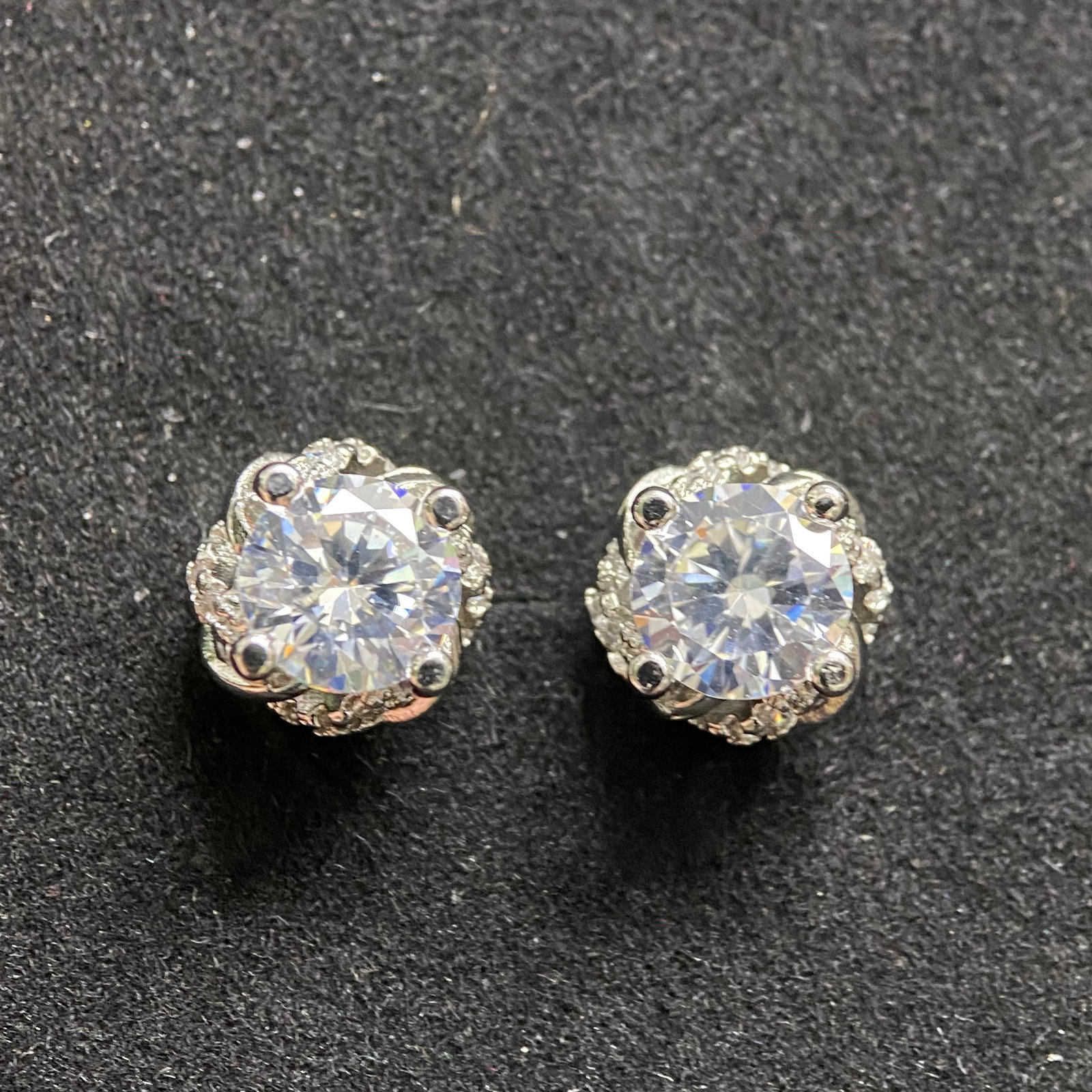 Classic Round-Cut Moissanite Stud Earrings: A pair of elegant stud earrings featuring brilliant round-cut moissanite gemstones. Each stone is held in a minimalist four-prong setting of polished silver-tone metal, designed to highlight the stone