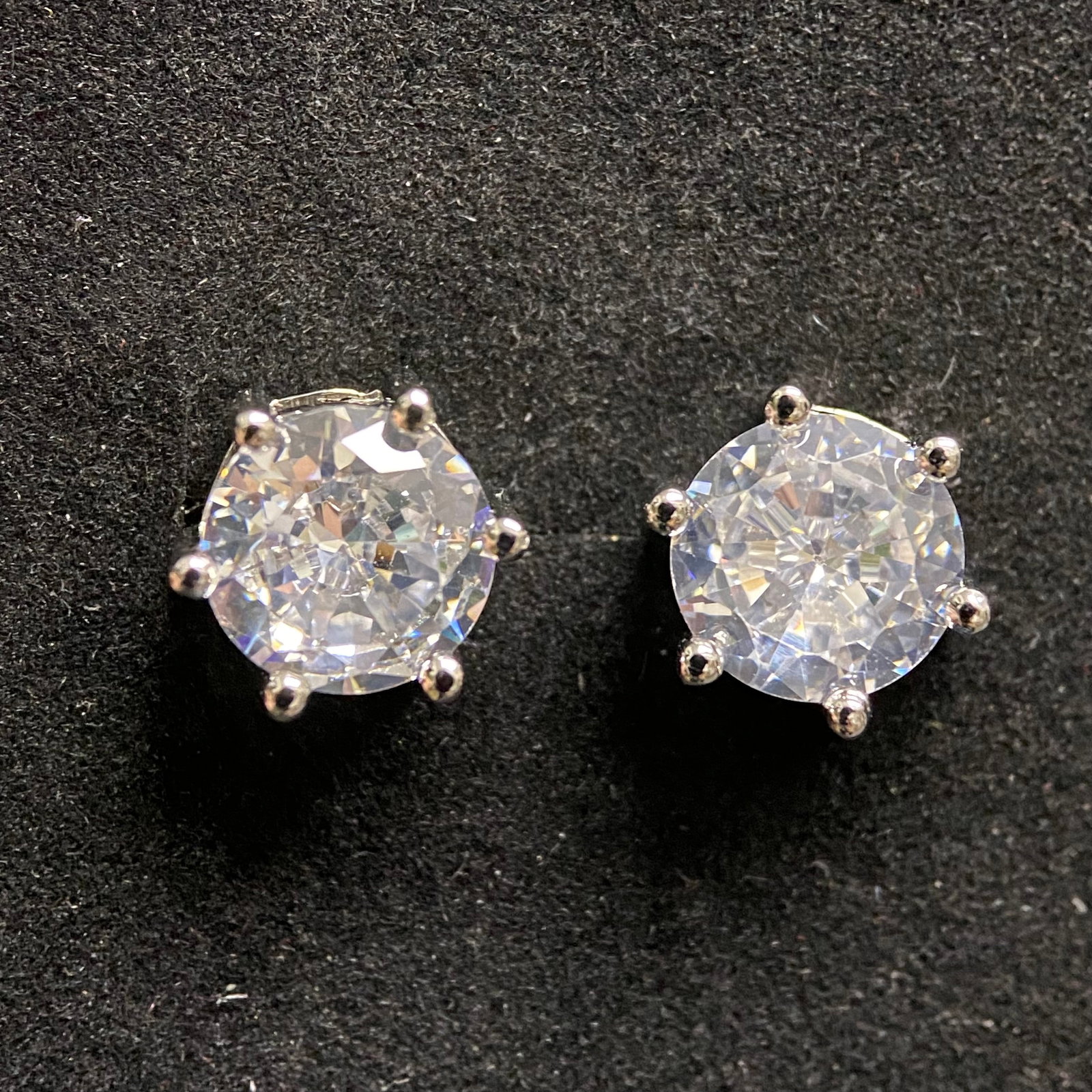 Classic Round-Cut Moissanite Stud Earrings: A pair of sophisticated stud earrings featuring brilliant round-cut moissanite gemstones. Each stone is secured by a classic four-prong basket setting in polished silver-tone metal, providing a timele