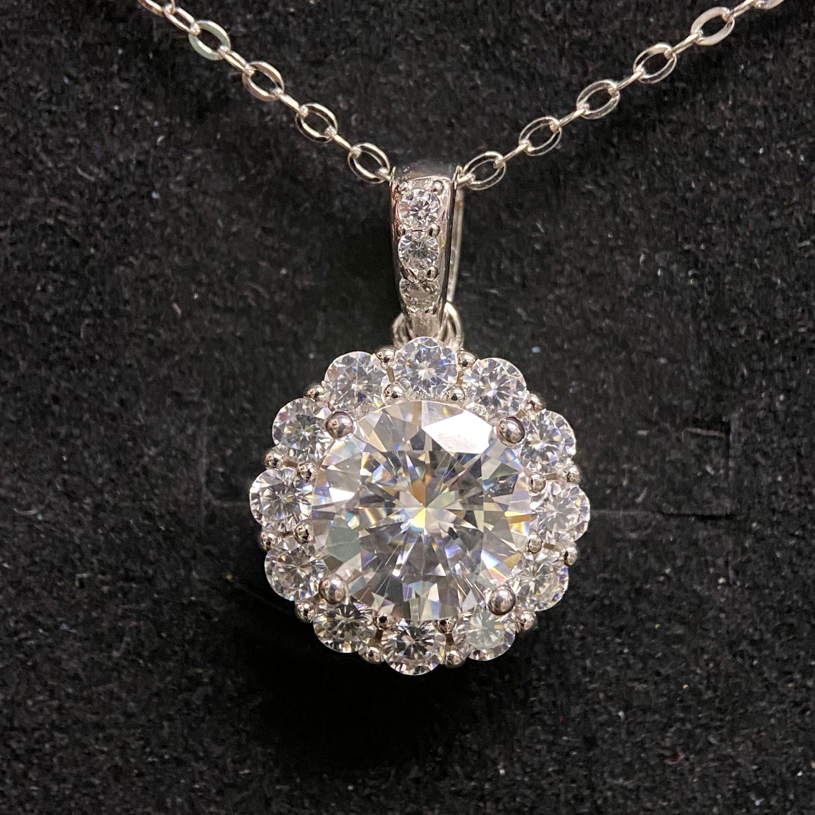 Round Brilliant Cut Halo Pendant: A classic and elegant white metal pendant featuring a prominent round brilliant-cut central stone. The center stone is bordered by a continuous halo of smaller round stones, adding significant brillia