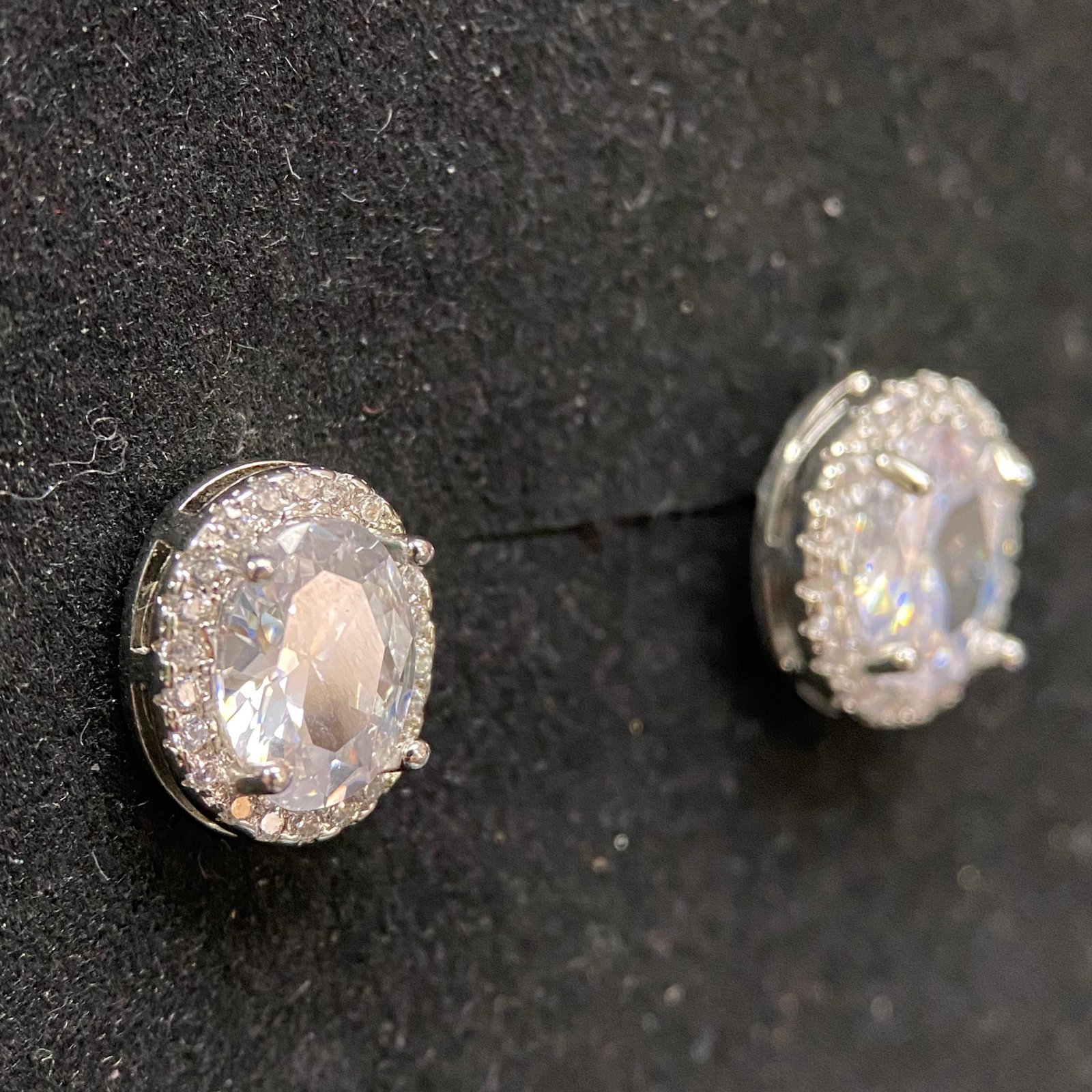 Moissanite Halo Earrings – 2.0ct D, 7x9mm Oval Cut – GRA Certified – Scalloped Pavé Border: This pair of exquisite stud earrings features two center-set oval cut stones secured by four polished prongs. Each center stone is encircled by a brilliant halo of pavé-set accents in a scalloped set