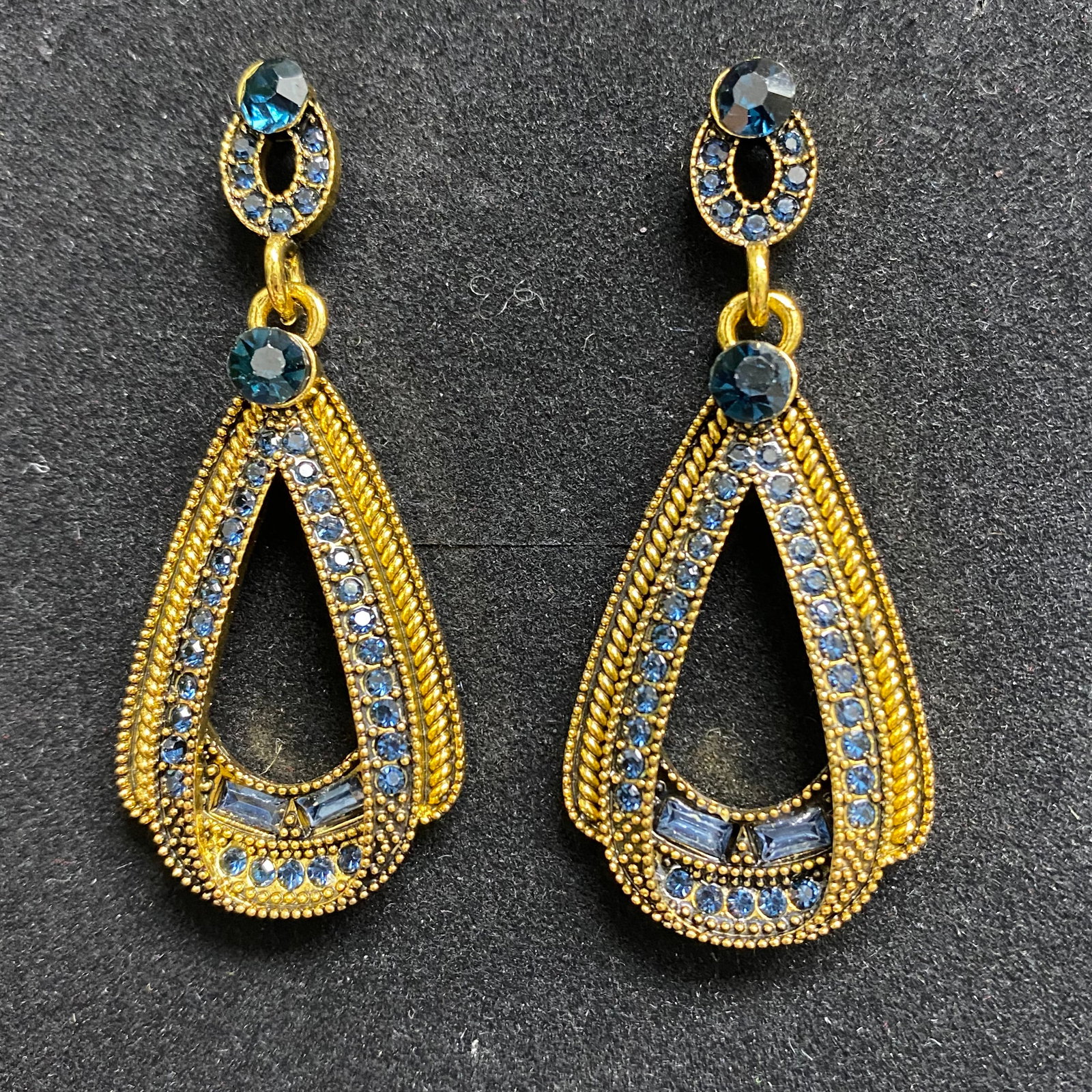 Vivid Blue Stone Halo Stud Earrings – Polished Silver-Toned Mounting: This pair of sophisticated stud earrings features two vibrant blue center stones in a classic round cut, each encircled by a delicate halo of brilliant colorless accents. The stones exhibit a deep, co