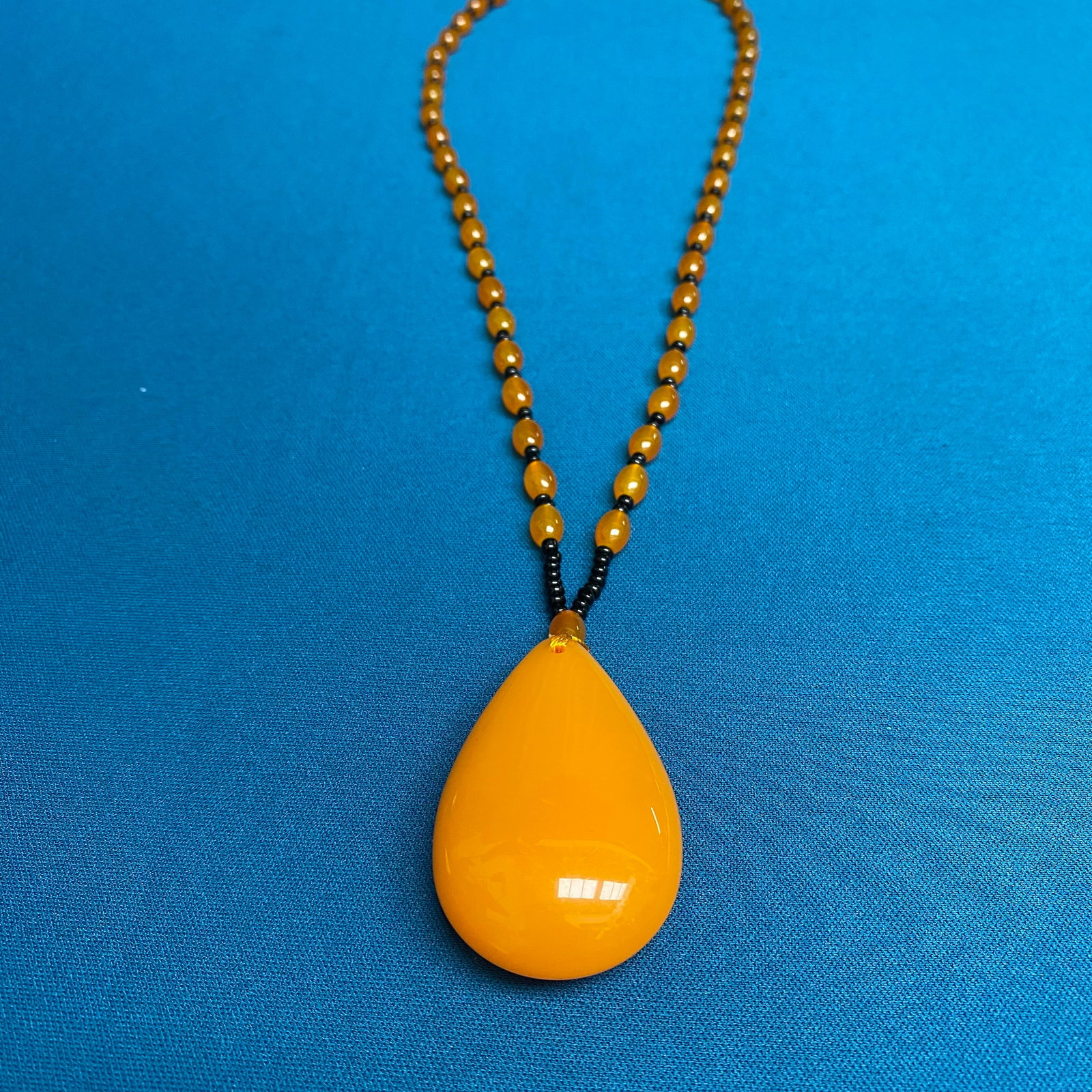 Oversized Amber-Tone Teardrop Pendant with Beaded Necklace: A bold and substantial statement piece, this necklace features a monumental teardrop-shaped pendant in a rich, opaque butterscotch amber hue. The pendant is expertly polished to a high-gloss, mirror-l