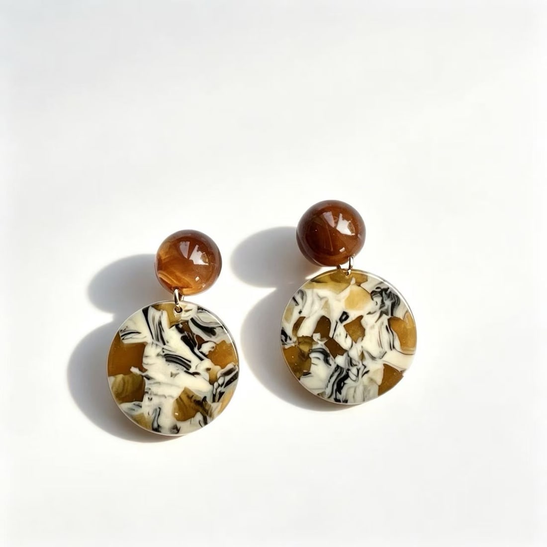 Amber Tone Resin Disc Dangle Earrings: Pair of contemporary dangle earrings featuring glossy amber-tone round studs with marbled resin disc drops in cream, brown, and black tones. Modern statement jewelry design.
