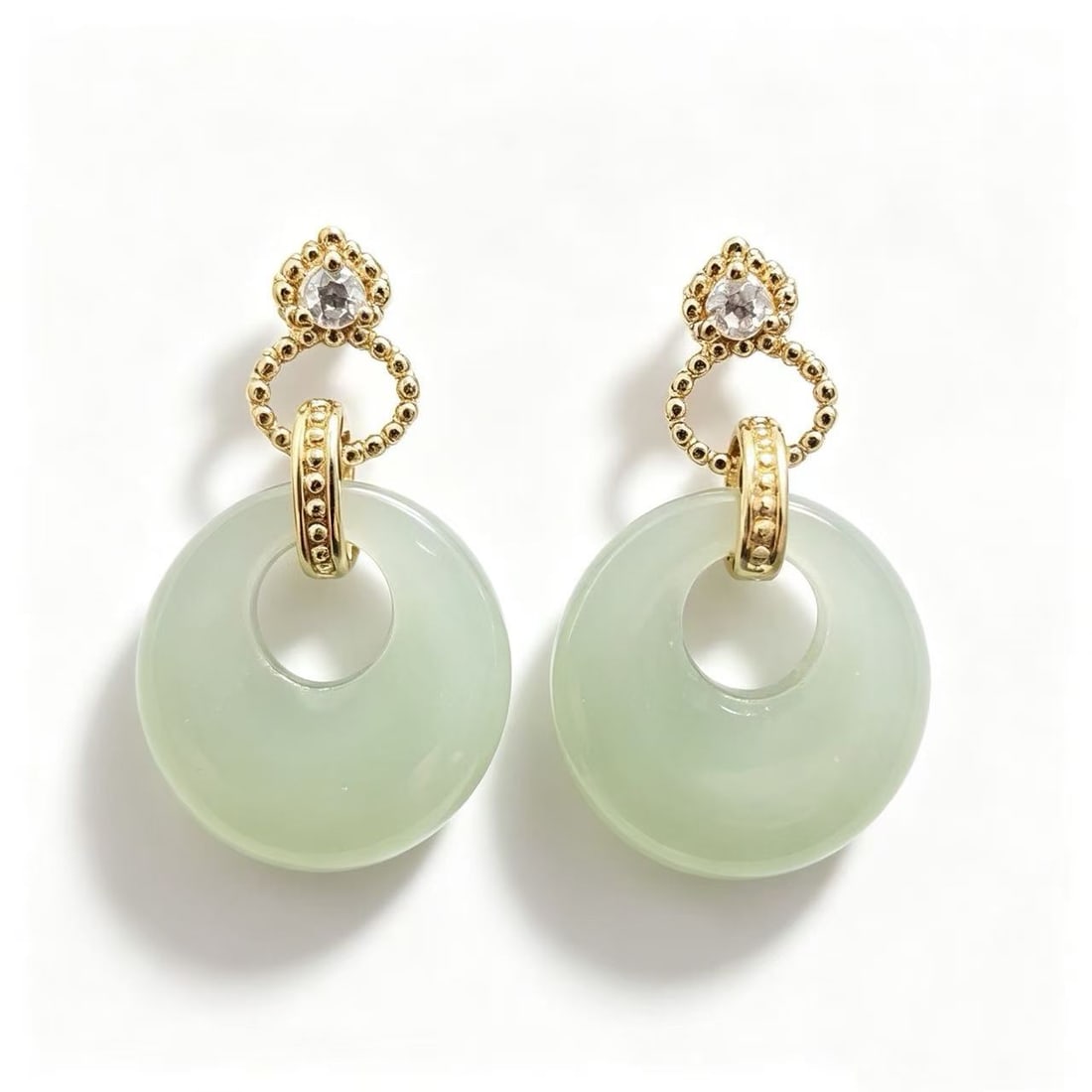 Gold-Gilded Jade Style Circular Drop Earrings: Pair of gold-gilded drop earrings featuring pale green jade-style circular disc elements suspended from decorative gold-tone settings with clear accent stones. Designed in a contemporary-inspired aest