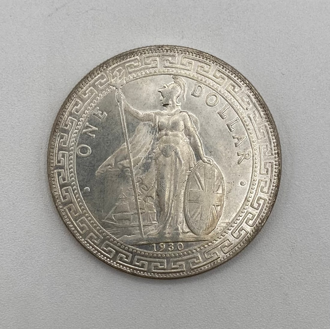 1930 British Trade Dollar One Dollar Silver Round / Coin: Design: Standing Britannia with trident and shield against a ship background, framed by a classic Greek key border motif, dated 1930. Denomination / Text: "ONE DOLLAR" Type: Silver Round / Trade Dolla