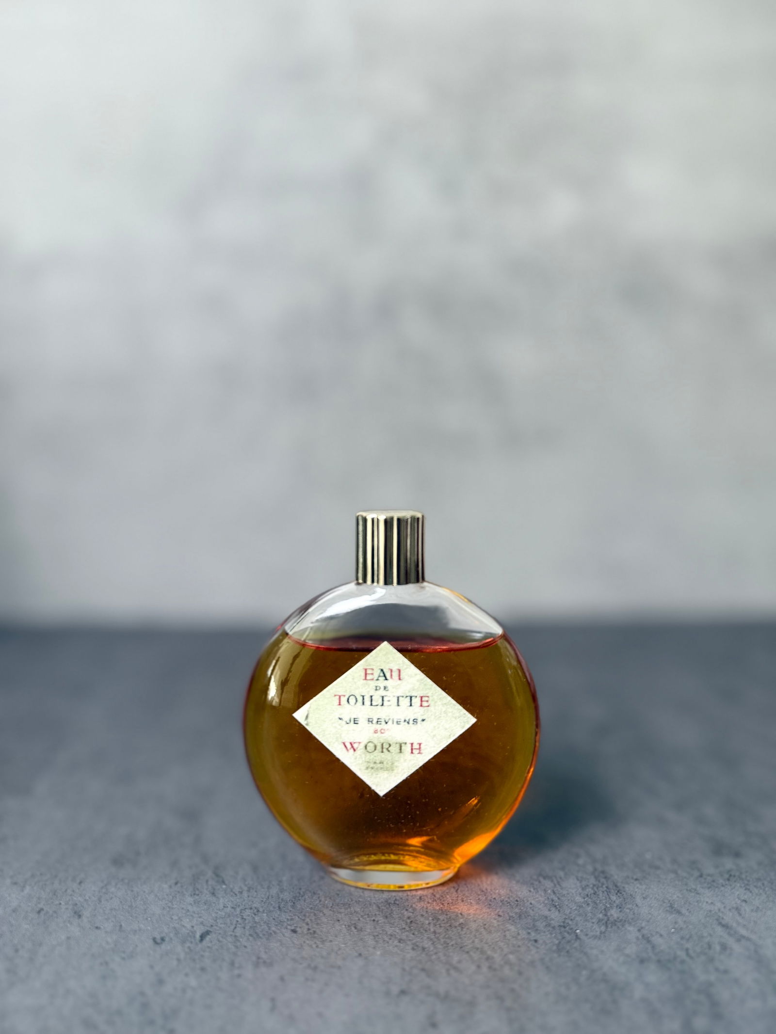 Vintage Worth “Je Reviens” Eau de Toilette Bottle: A beautifully preserved vintage bottle of Worth “Je Reviens” Eau de Toilette, presented in a rounded amber-toned glass form with its original label intact. The warm golden hue of the liquid