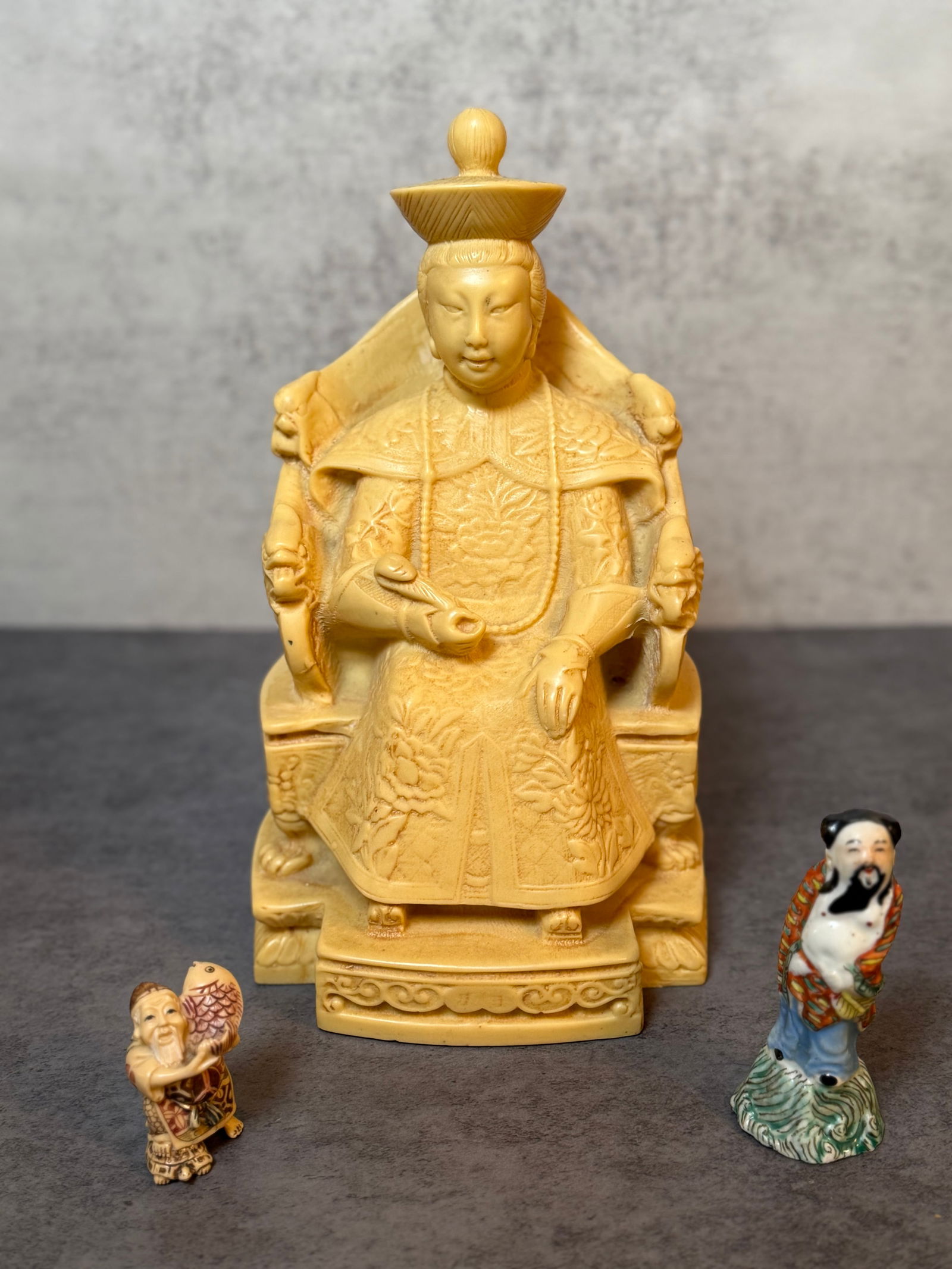 Asian Style Carved Seated Emperor Figure, Decorative Resin Composition, with Two Miniature Figural: This Asian style carved figure depicts a seated imperial dignitary in formal ceremonial attire, presented upon an ornate throne with attendant mythical beast-form arm supports.The figure is finely