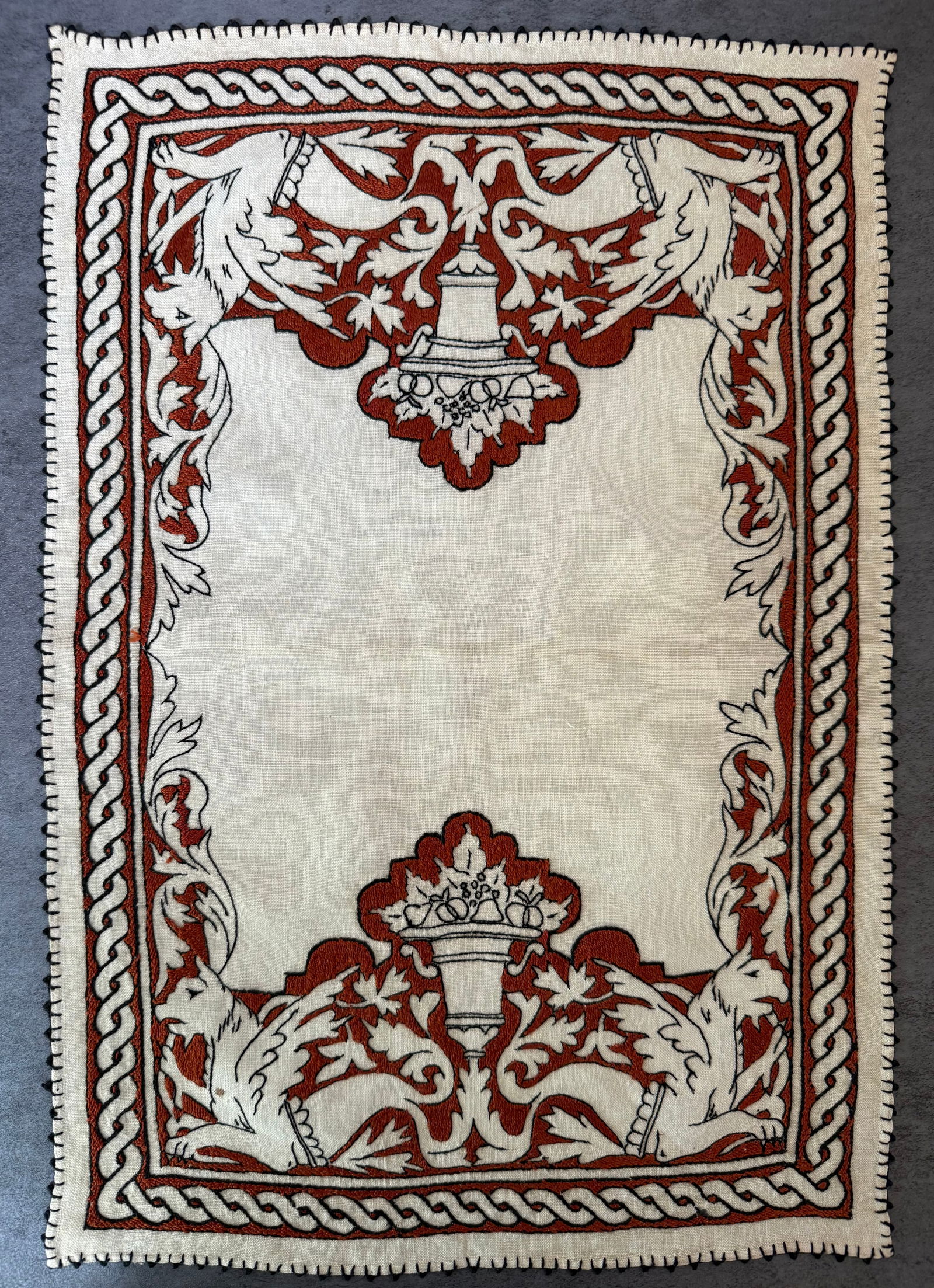 Vintage Hand Embroidered Linen Table Mat with Heraldic Griffin Motif: A vintage hand-embroidered linen table mat featuring an elaborate decorative border with heraldic griffin figures and classical urn motifs. The embroidery is worked in red thread on natural linen, cre