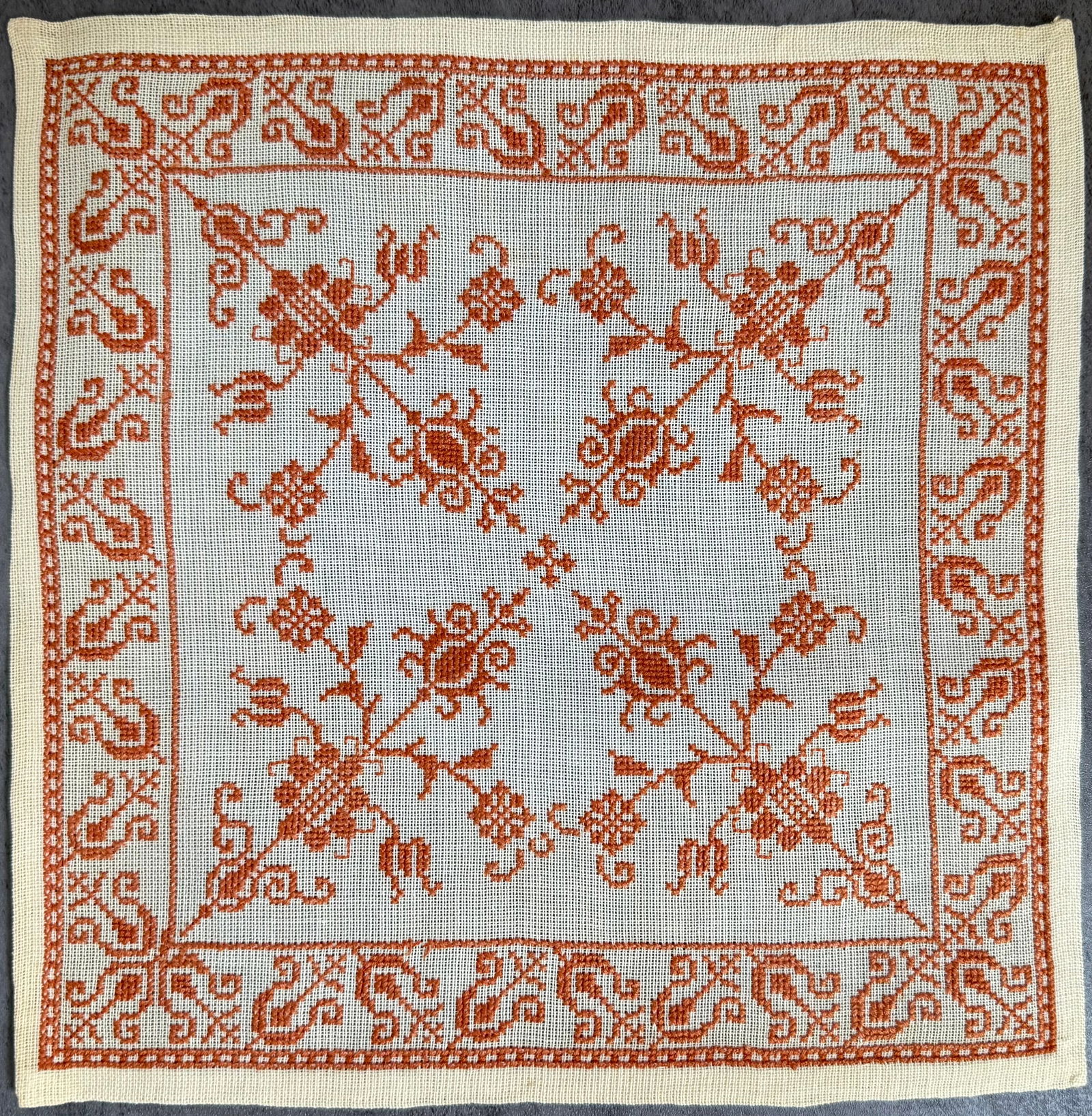 Vintage Hand Embroidered Linen Textile Panel with Floral Folk Motif: A vintage hand-embroidered linen textile panel featuring a symmetrical floral folk motif in rust-colored thread. The design displays scrolling floral elements arranged in a circular composition,