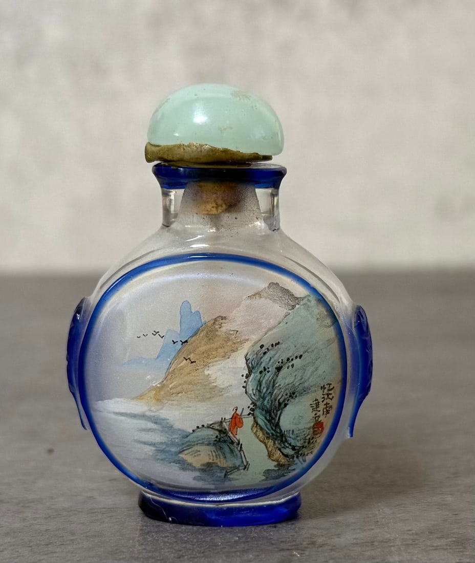 Chinese Blue Overlay Inside Painted Glass Snuff Bottle: Chinese inside-painted glass snuff bottle of rounded form with blue overlay decoration framing the central panels and molded mask-form handles at the sides. One side is decorated with a blooming