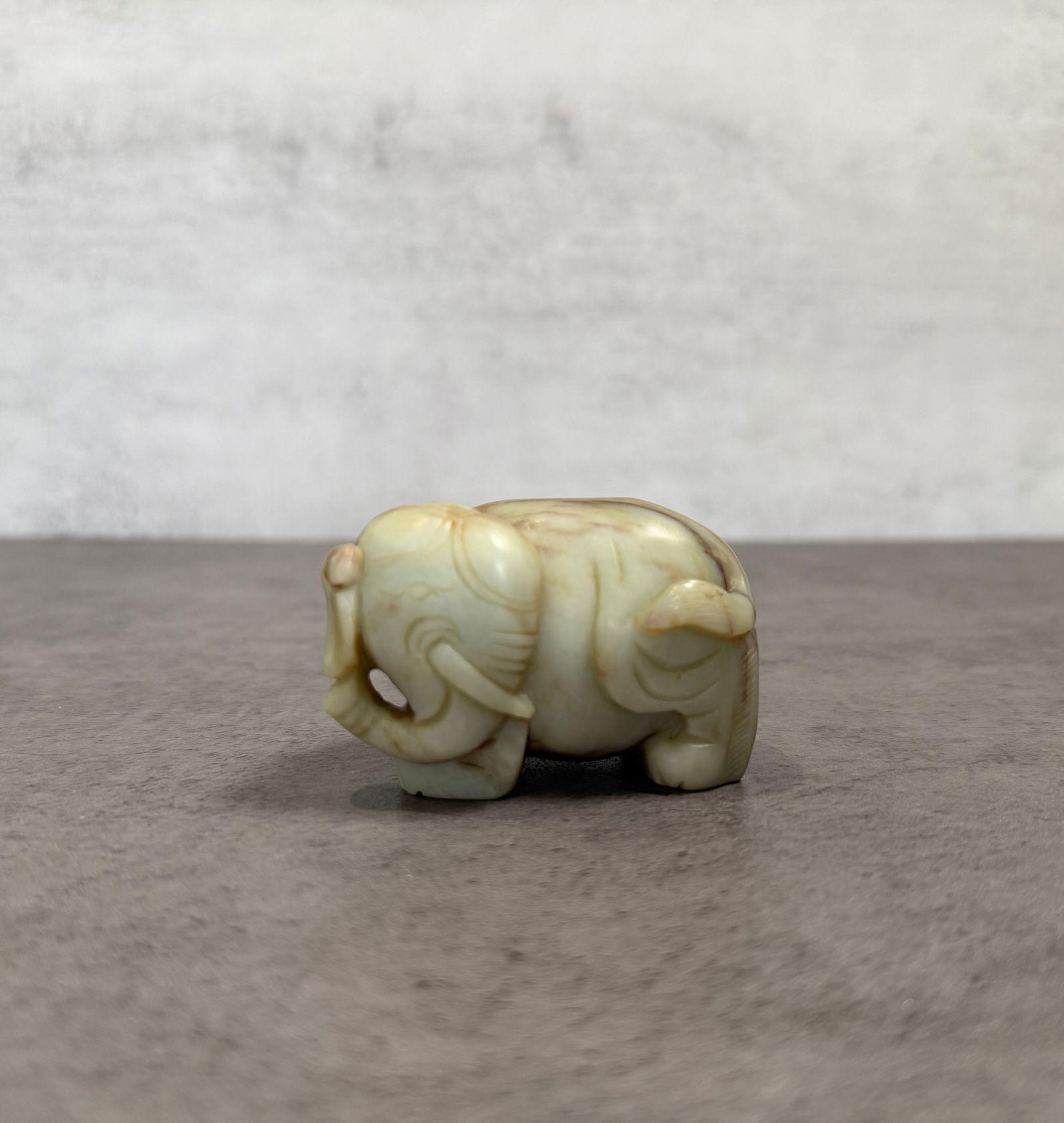 Chinese Carved Jadeite Elephant Figurine: A Chinese carved jade elephant figurine formed in a compact standing posture. The sculpture displays softly rounded carving with incised decorative lines along the body and natural stone coloration
