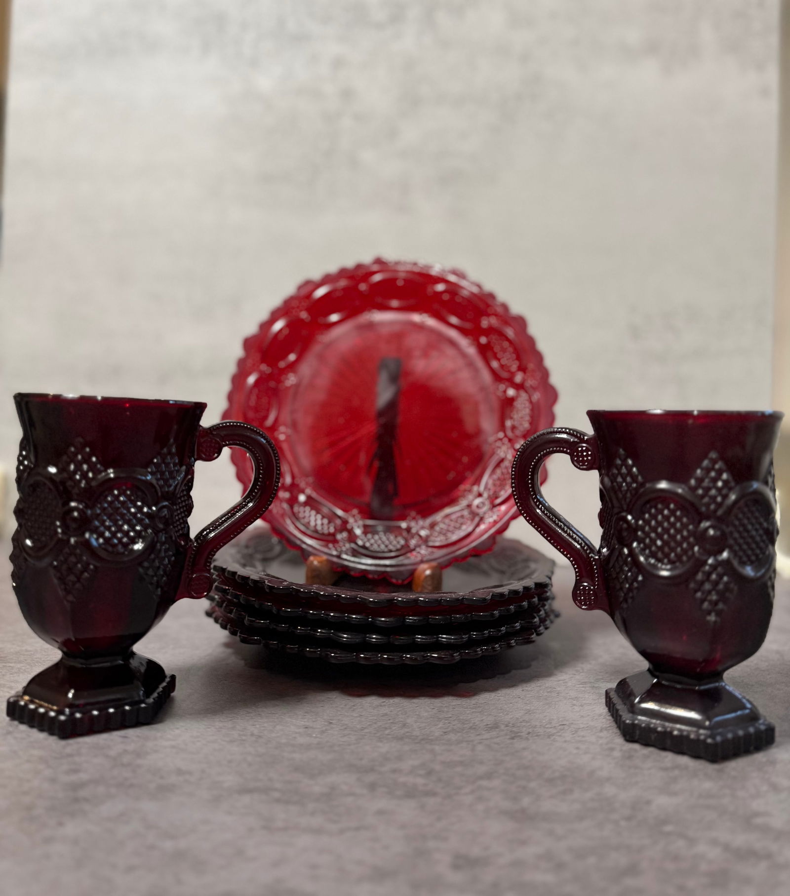 Avon Cape Cod Ruby Red Glass Cups and Saucers Set, Vintage Pressed Glass Tableware: A classic Avon Cape Cod collection set in deep ruby red pressed glass, featuring handled cups accompanied by matching saucers.The pieces display Avon’s iconic Cape Cod pattern, characterized by