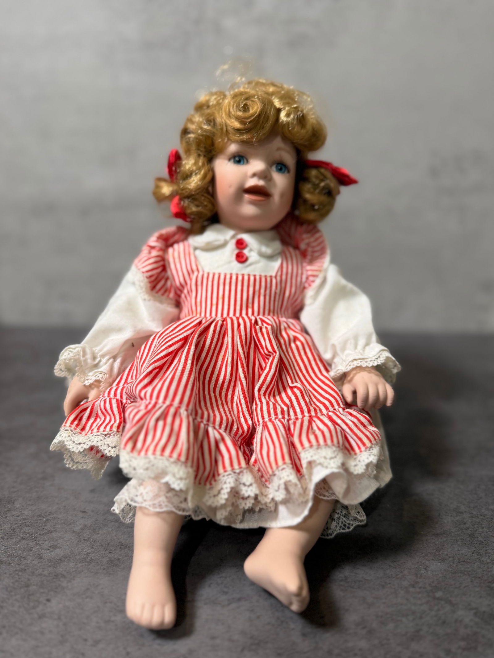 Vintage Porcelain Doll with Curly Hair, Blue Eyes, Red & White Dress, Collectible Decorative Figure: A charming vintage porcelain doll featuring expressive blue eyes, delicately painted facial details, and curly blonde hair styled with red ribbon accents.She is dressed in a red and white striped