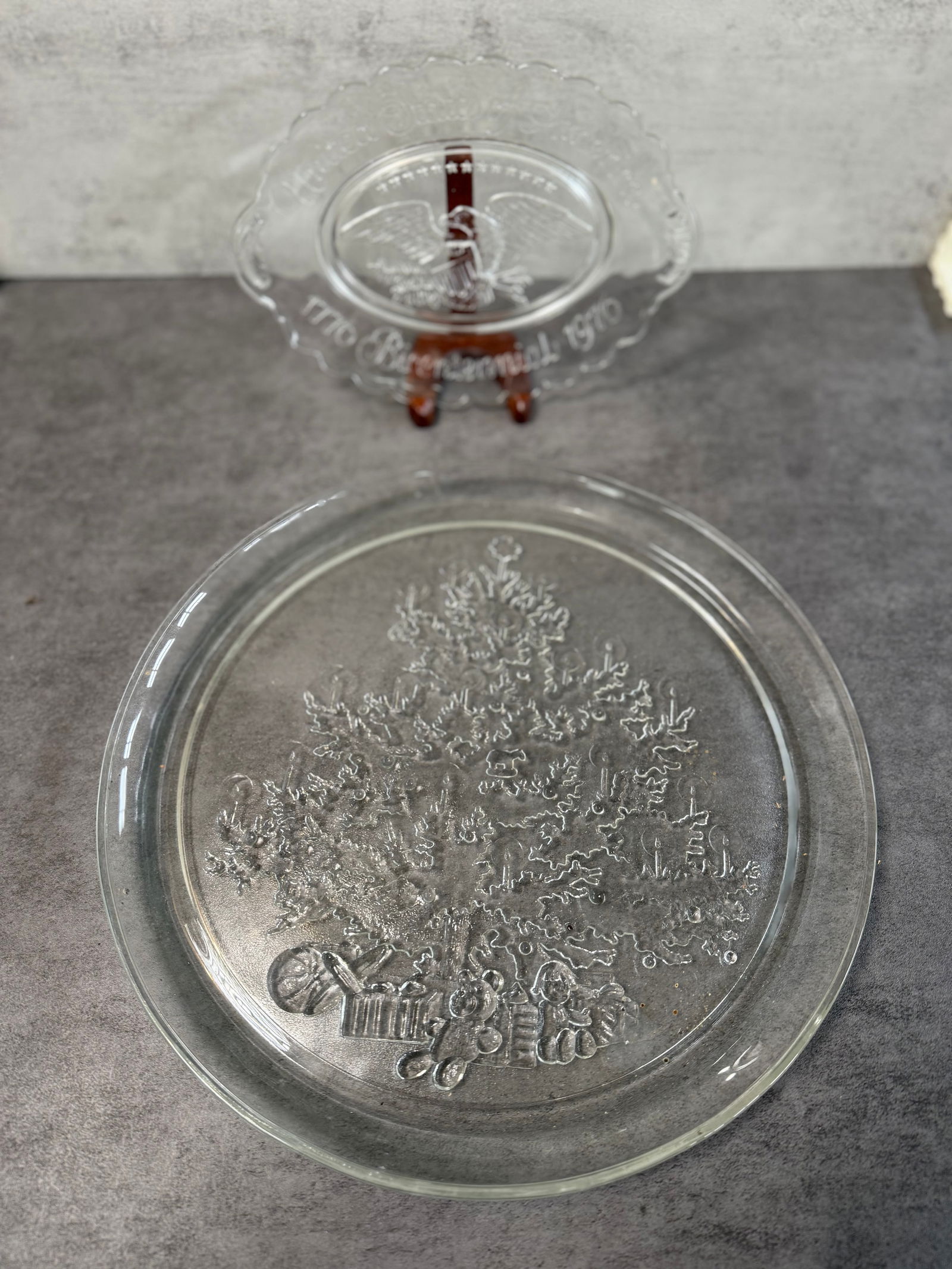 Vintage Pressed Glass Plates, Christmas Tree & 1976 Bicentennial Eagle (2 pcs): A pair of vintage pressed glass plates, including a holiday-themed Christmas tree design and a commemorative 1976 Bicentennial plate featuring an eagle motif.Both plates are molded in clear glass