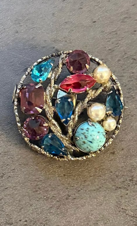 Vintage Multi-Stone Brooch with Colored Glass, Faux Pearl and Gold Tone Setting: A vintage brooch featuring a vibrant arrangement of multicolored stones set within a textured gold tone framework. The composition includes faceted and cabochon-style glass elements in shades of