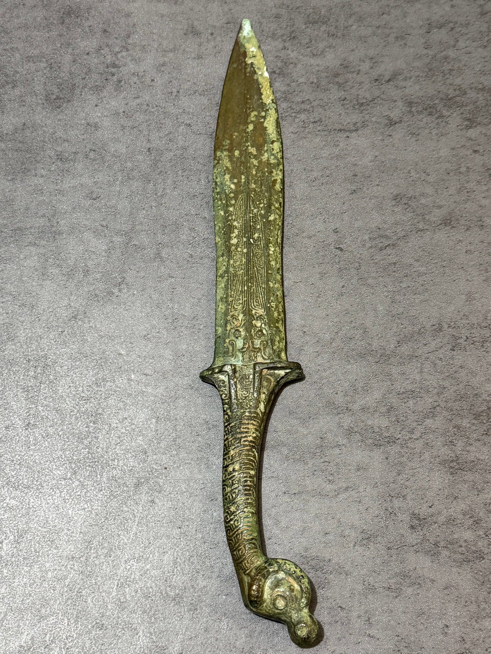 Asian Bronze Ceremonial Dagger With Zoomorphic Handle: Asian bronze ritual dagger featuring an elongated blade with finely incised geometric and stylized motifs throughout. The handle is cast with textured ornamentation and terminates in a sculptural