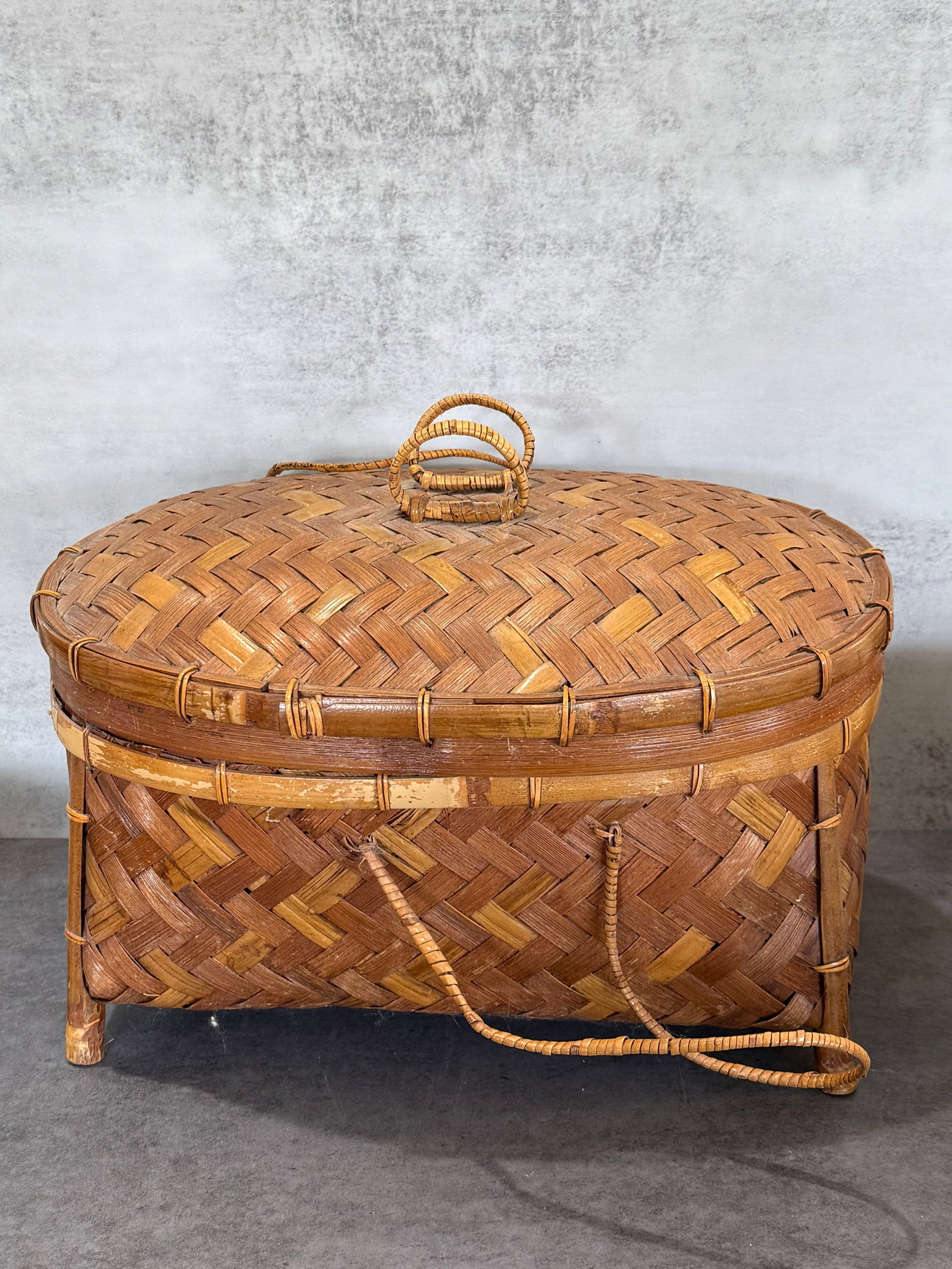 Handwoven Bamboo Basket Box with Raised Bamboo Legs and Cover: A vintage handwoven bamboo storage basket of oval form, featuring an intricate woven pattern in contrasting natural bamboo tones. The basket is raised on distinctive bamboo-form legs and fitted with
