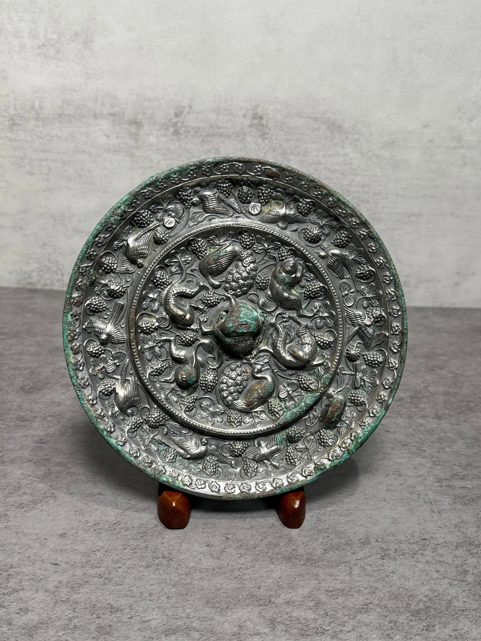 Chinese Silvered Bronze Relief Mirror with Birds & Fruit Motifs: Chinese silvered bronze mirror featuring densely layered relief decoration of birds, grapes, peaches, and scrolling vines surrounding a raised central medallion. The richly worked surface and archaist