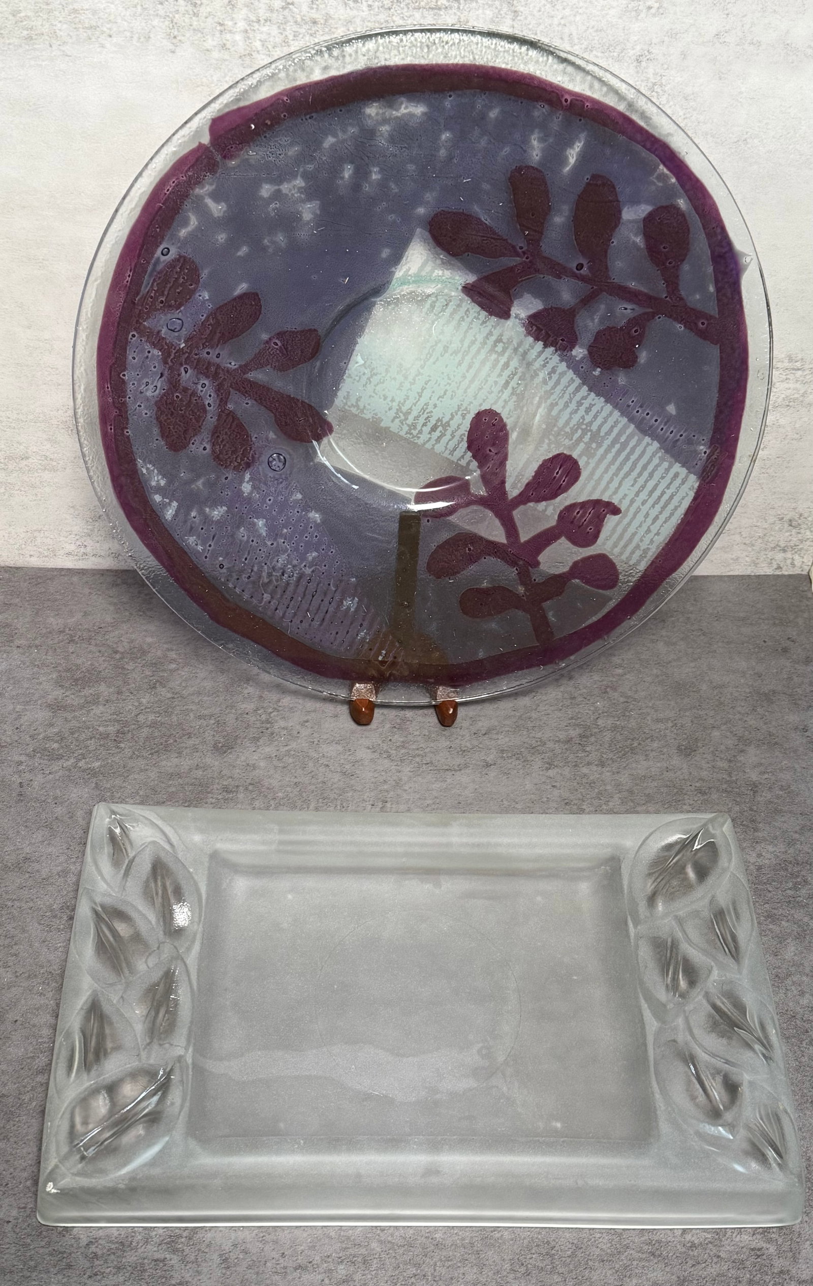 GROUP OF LARGE ART GLASS SERVING TRAYS: A group of two art glass serving trays, including a circular example with deep amethyst-toned coloration and stylized botanical motifs, together with a frosted rectangular tray featuring molded