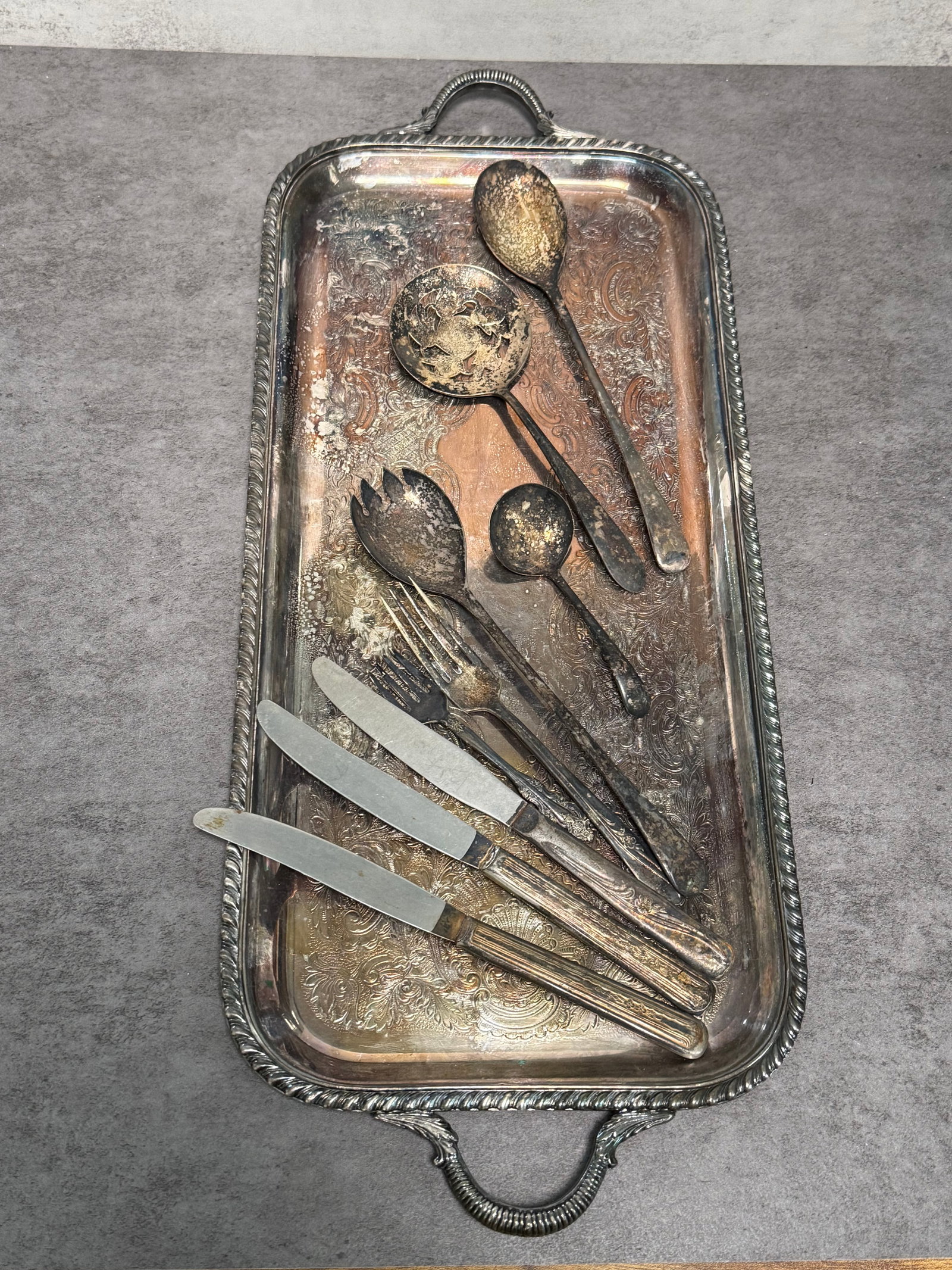 SILVER-PLATED SERVING TRAY WITH ASSORTED FLATWARE: A silver-plated serving group including an engraved handled tray together with assorted serving utensils and flatware, comprising ladles, serving forks, spoons, and knives. The tray features an ornate