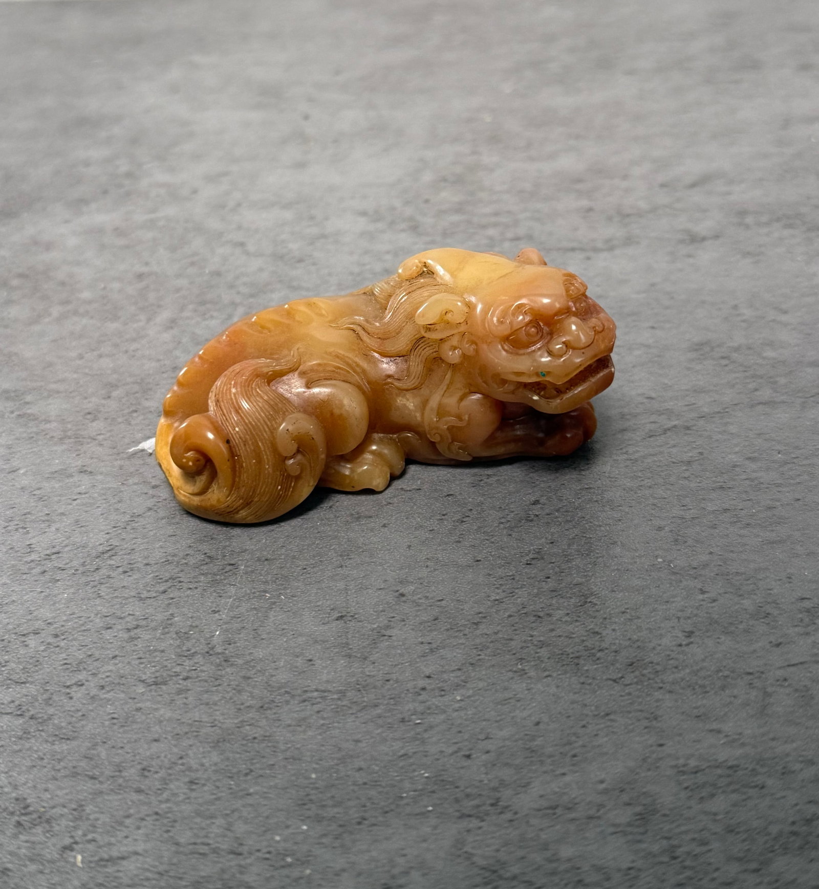 SHOUSHAN STONE CARVED MYTHICAL BEAST FIGURE: A finely carved Shoushan stone figure depicting a recumbent mythical beast with expressive facial features, scrolling tail, and flowing mane details. The stone displays warm honey and russet-toned