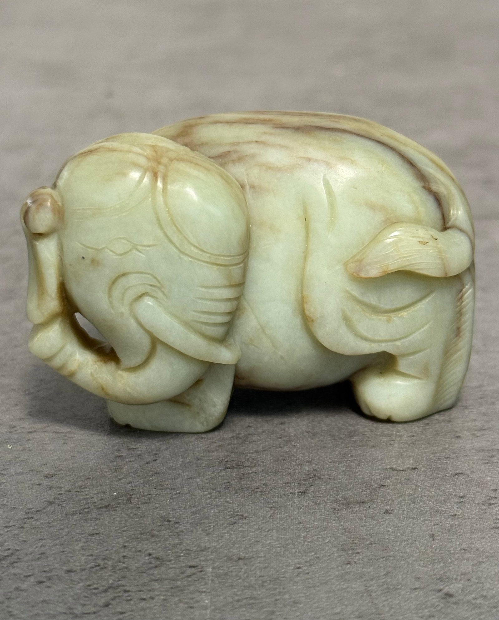 CHINESE JADEITE CARVED ELEPHANT FIGURE: A Chinese jadeite carved elephant figure depicted in recumbent form with curled trunk and stylized incised detailing throughout. The stone displays soft celadon coloration with natural russet