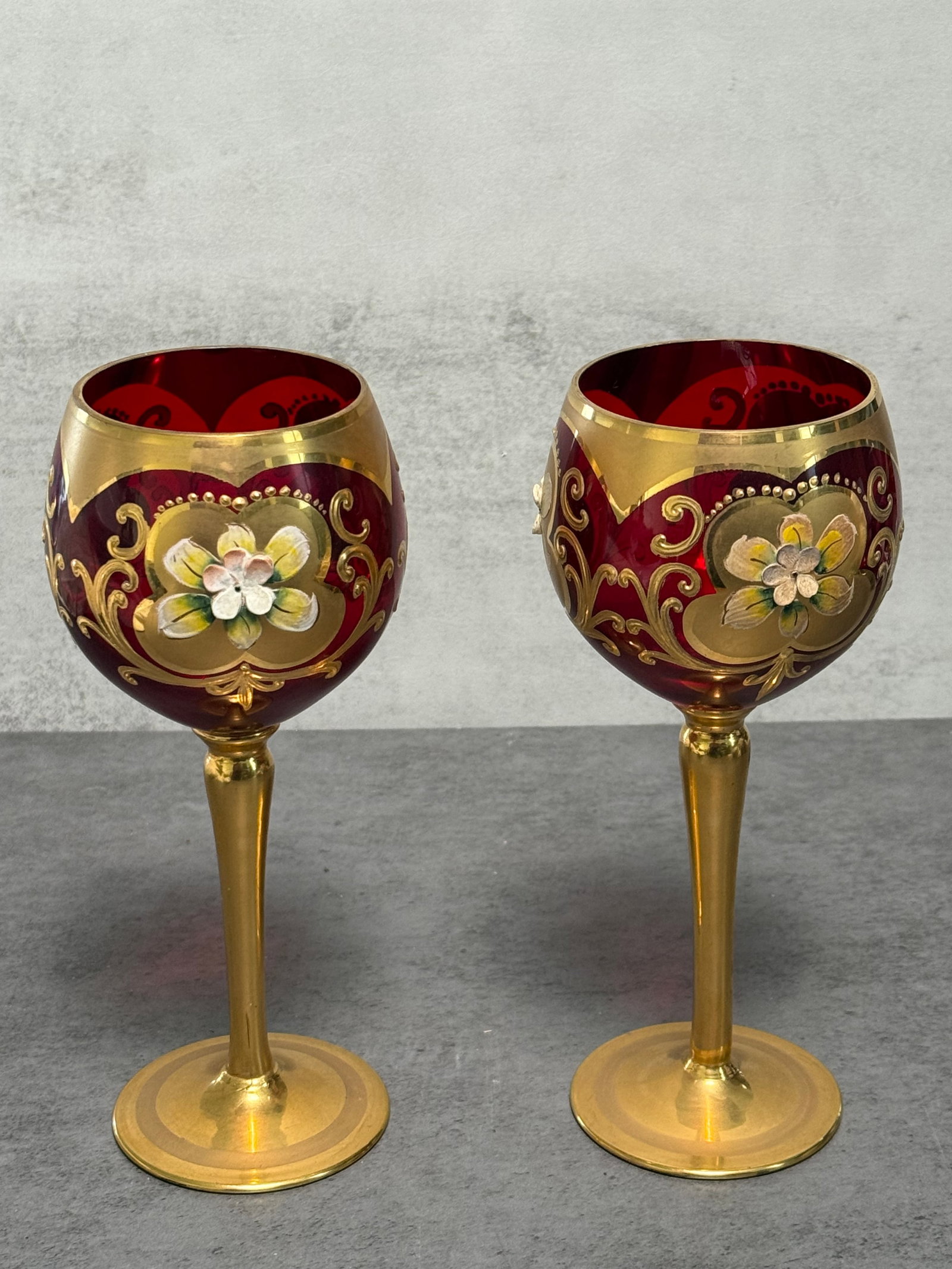 Vintage 2 T.M.J. Wine Glasses 24K Gold Leaf - Red: A pair of vintage T.M.J. wine goblets in deep ruby red glass, accented with 24K gold decoration and hand-painted floral motifs. The bowls are embellished with scrolling gilt ornamentation and