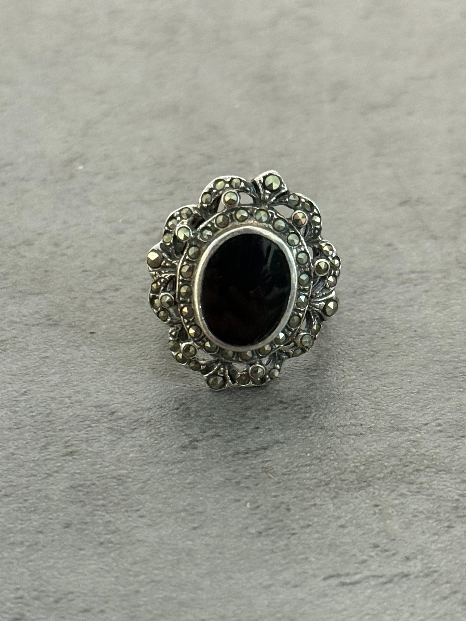 VINTAGE STERLING SILVER, ONYX & MARCASITE RING: A vintage sterling silver ring featuring an oval black onyx cabochon surrounded by marcasite stones in an ornate openwork setting. The design displays elegant floral and scroll-form detailing