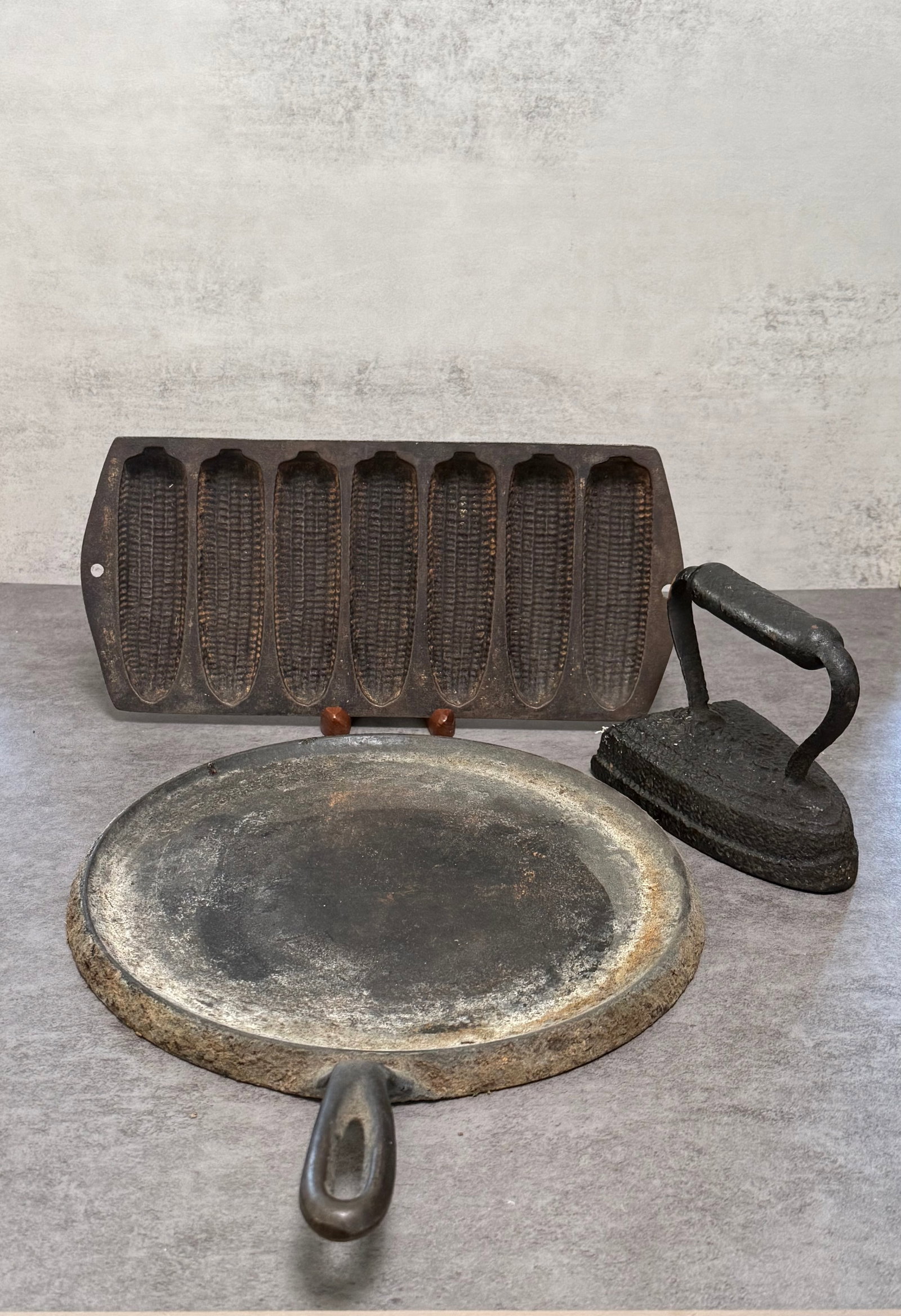 Group Of Cast Iron ITEMS: Group of antique cast iron kitchenware items including a corn stick baking mold, flat iron, and handled griddle or skillet plate. Featuring heavily aged surfaces and rustic patina throughout, these