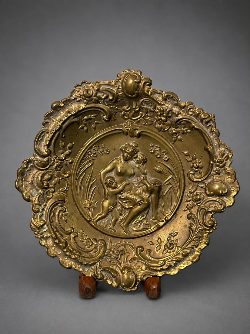 Victorian Style Repoussé Brass Decorative Tray with Classical Figural Scene, 8 Inch Diameter: An ornate Victorian-style brass decorative tray featuring an embossed classical figural scene of a seated female with cherubs, surrounded by an elaborate Rococo-inspired border of scrolling foliage an