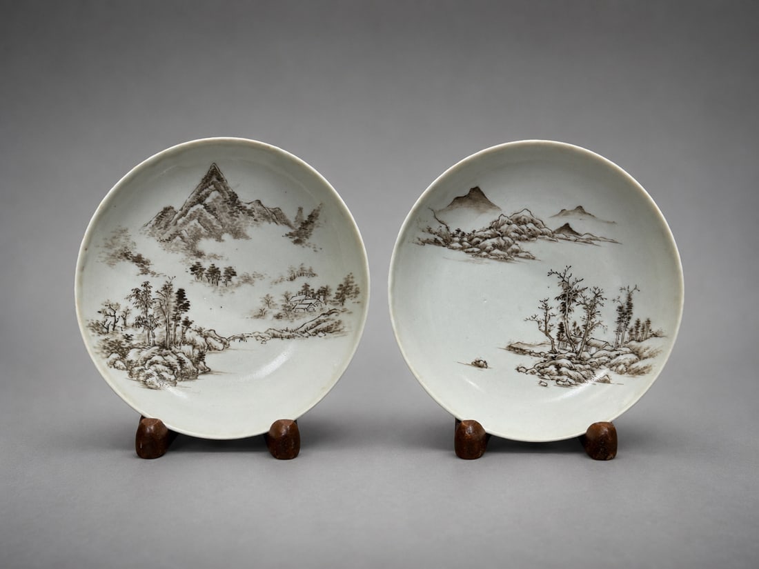 Pair of Chinese Grisaille Landscape Porcelain Dishes: A pair of Chinese porcelain dishes decorated in grisaille with finely painted landscape scenes depicting mountains, trees, riverside dwellings, bridges, and rocky terrain. Each dish features a