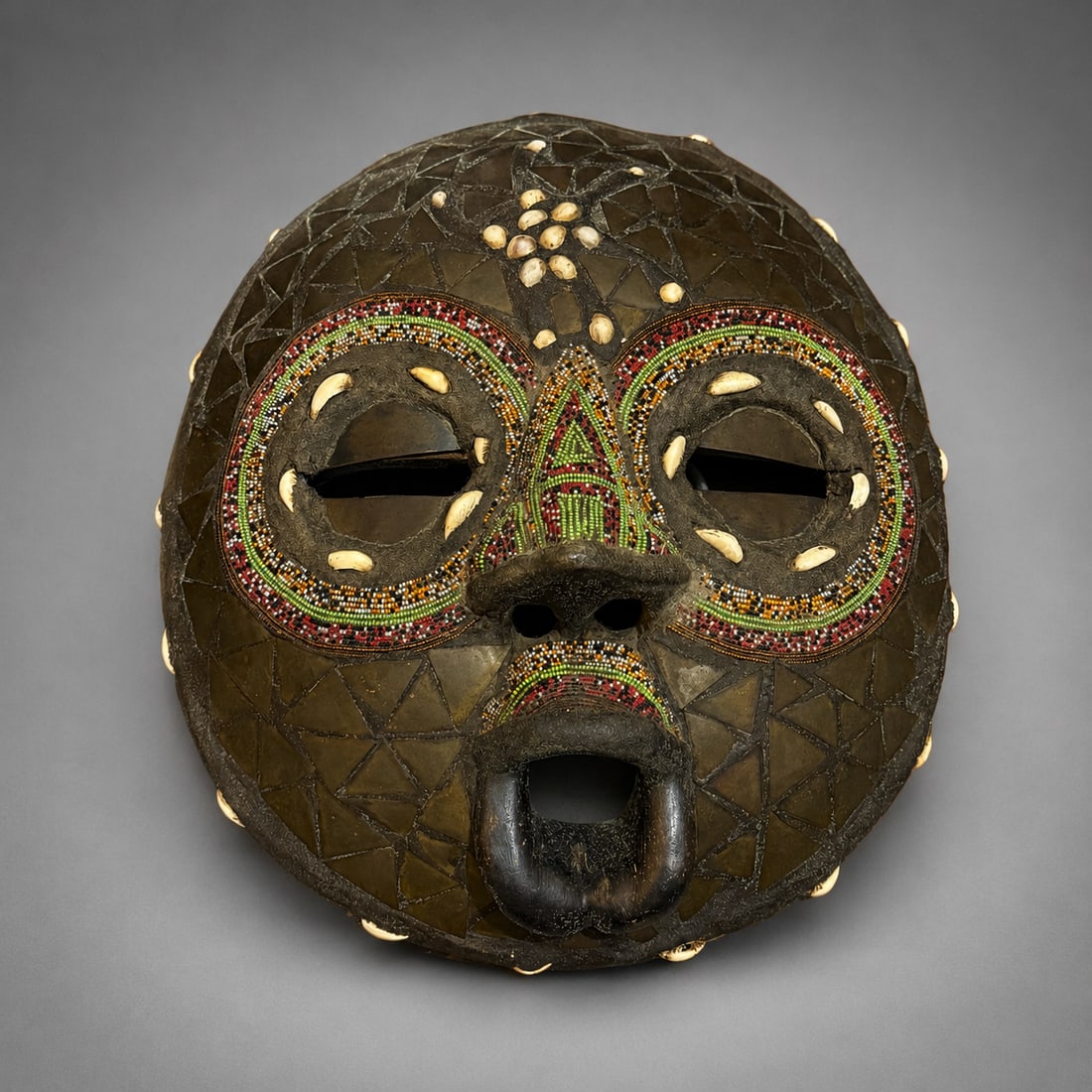 African Beaded and Shell-Inlaid Tribal Mask: An African tribal mask featuring intricate multicolored beadwork and shell inlay. The face is decorated with geometric applied panels, concentric beaded designs surrounding the eyes, shell accents, an