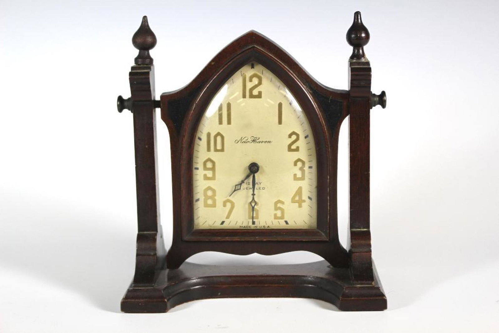 1930s New Haven 12 Day Jeweled Clock: A 1930s New Haven 12 Day Jeweled clock. This mantel clock features a wood frame with two carved posts with a finial resting on a tapered base. The clock rests between the two posts on a swivel post fe